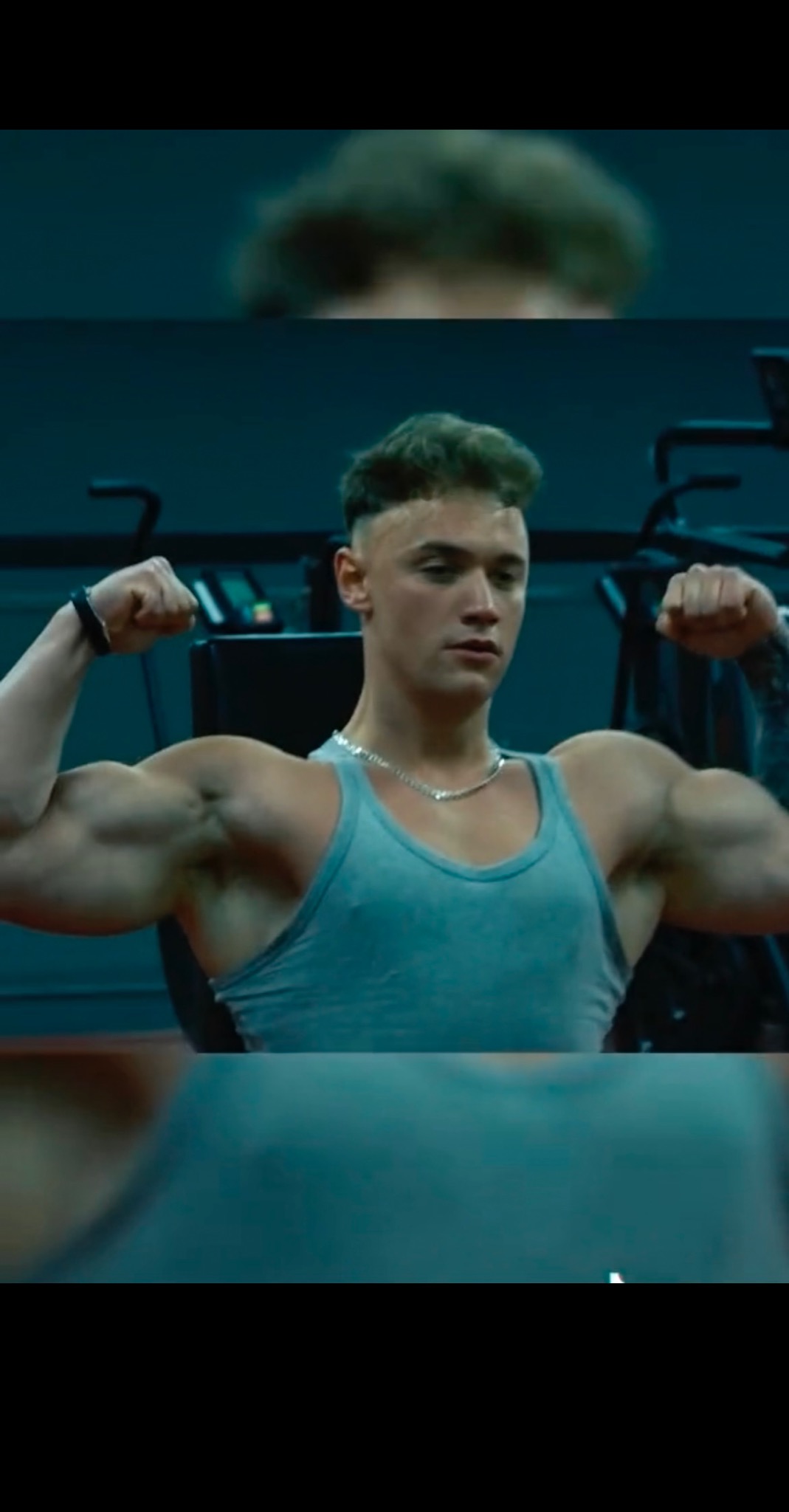 Młody mężczyzna w szarym podkoszulku prezentuje umięśnione bicepsy w siłowni, z delikatnym tatuażem widocznym na ramieniu i srebrnym łańcuszkiem na szyi.