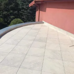 Wykończony taras na dachu budynku z jasnoszarych płyt betonowych, z widocznym obrzeżem z blachy i klimatyzatorem przy ścianie budynku.