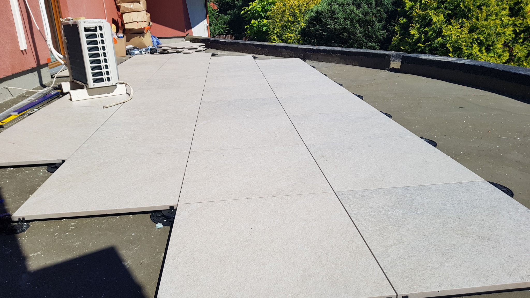 Montaż jasnych, kwadratowych płyt na regulowanych wspornikach na tarasie, widoczny klimatyzator i zieleń w tle.