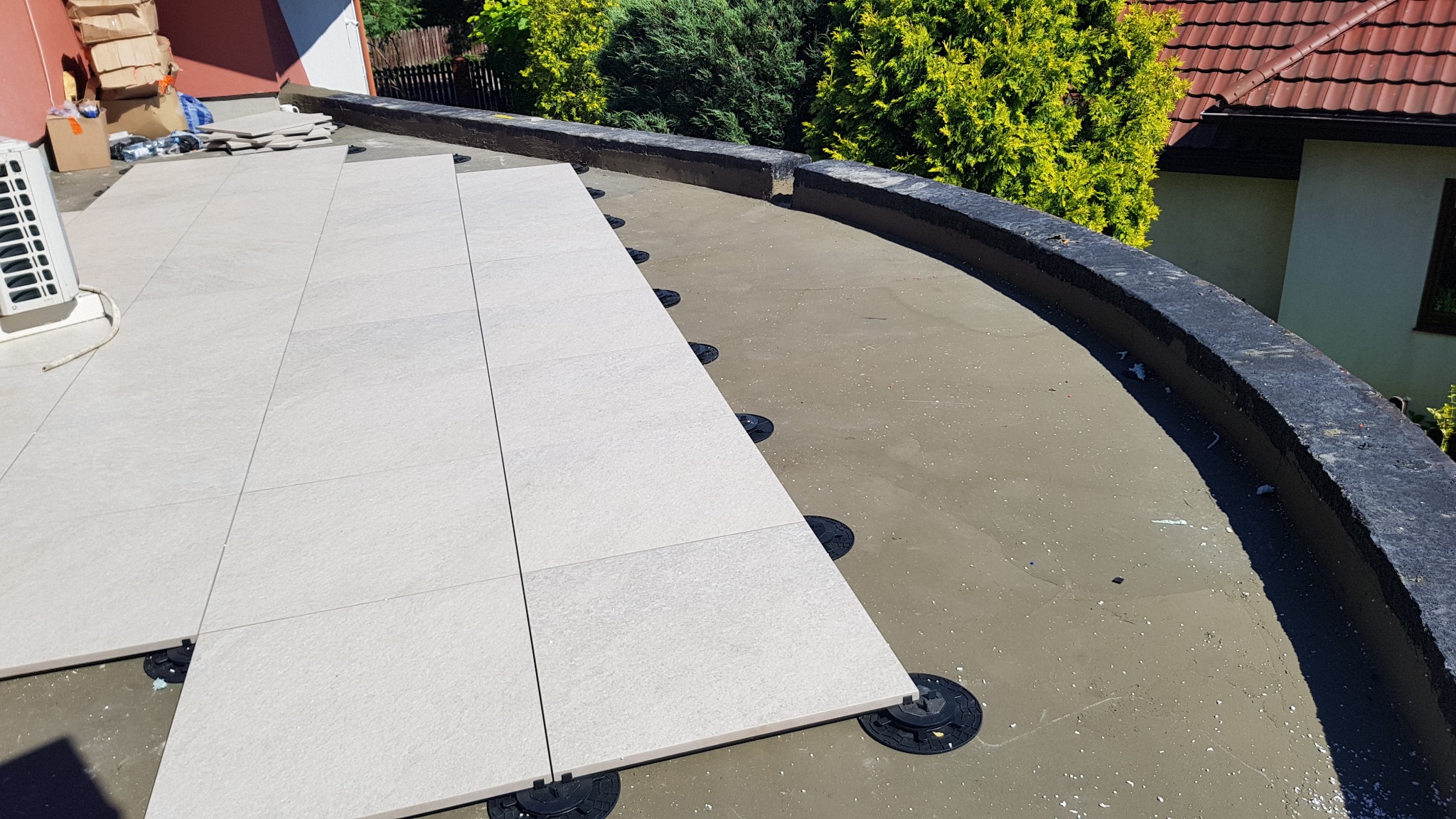 Układanie jasnych, kwadratowych płyt na regulowanych wspornikach na powierzchni tarasu, widoczny fragment instalacji klimatyzacji i elementy wykończeniowe dachu.