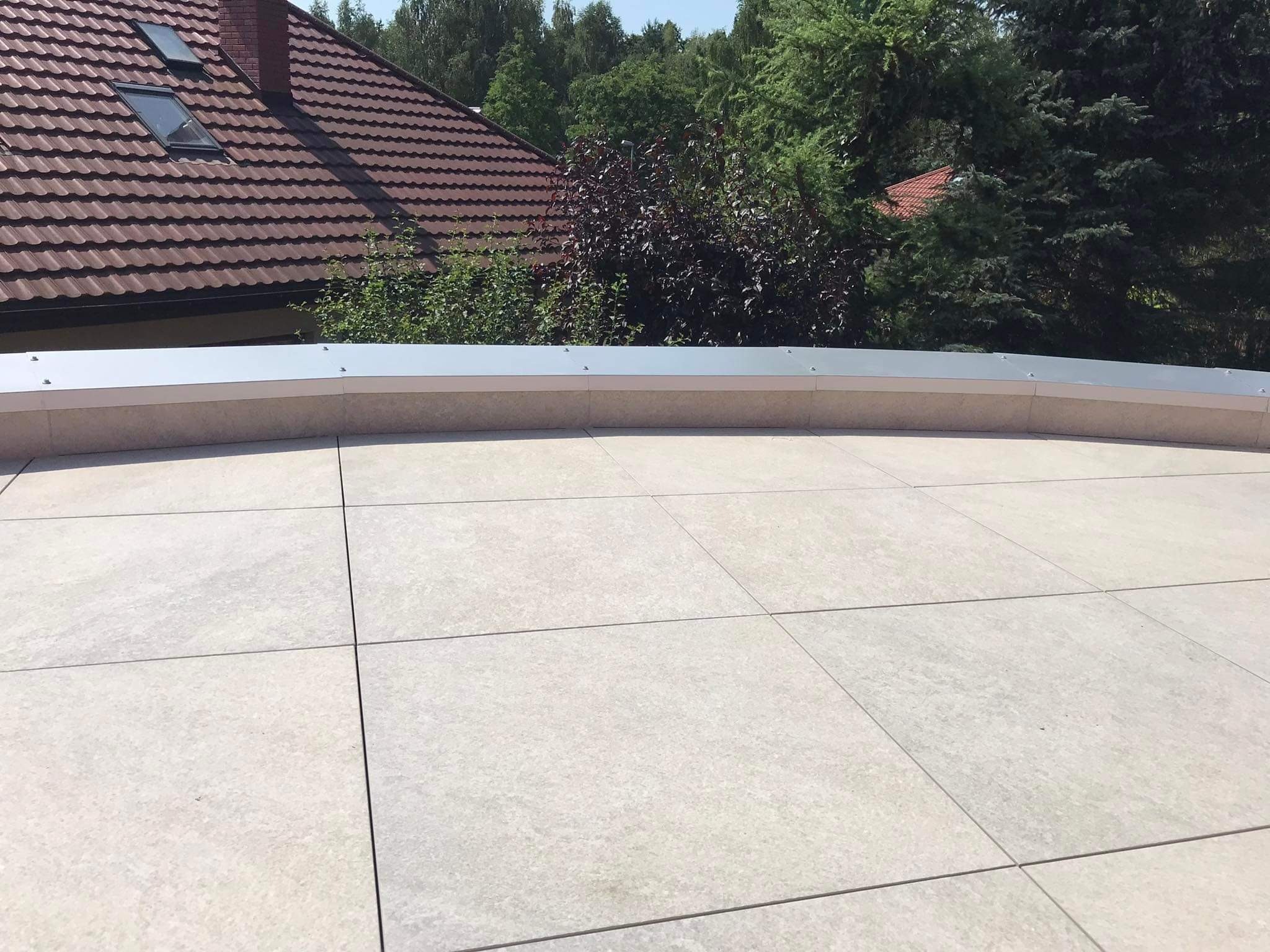 Wykończony taras z jasnych, kwadratowych płyt z widoczną metalową listwą krawędziową i fragmentem dachu domu w tle, otoczony zielenią.