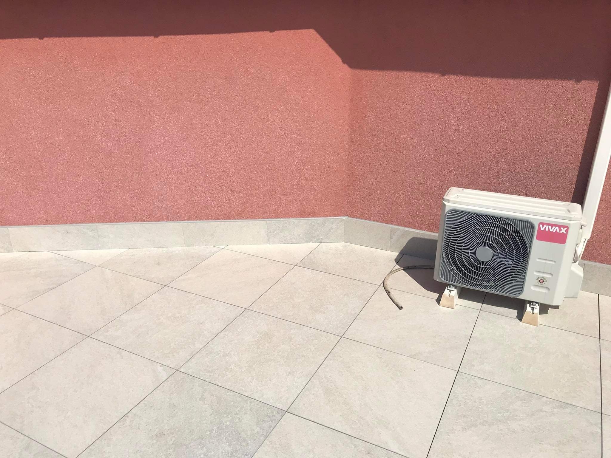 Zewnętrzna jednostka klimatyzacji marki Vivax zamontowana na jasnym, wykafelkowanym tarasie z różową ścianą w tle.