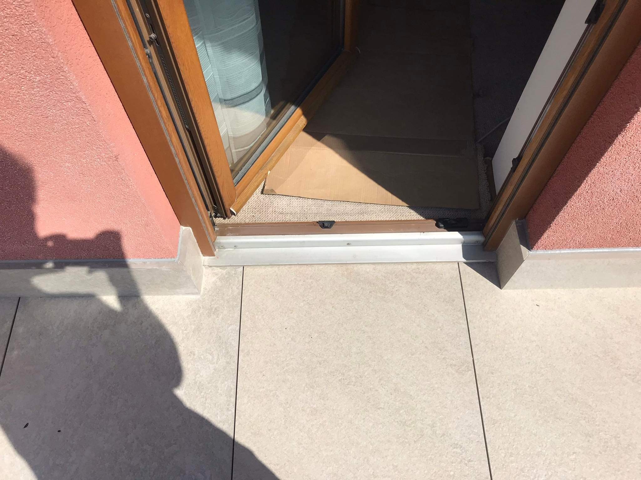 Położona jasna, kwadratowa terakota na tarasie, widok na próg drzwi balkonowych z brązową ramą i fragmentem różowej ściany budynku.