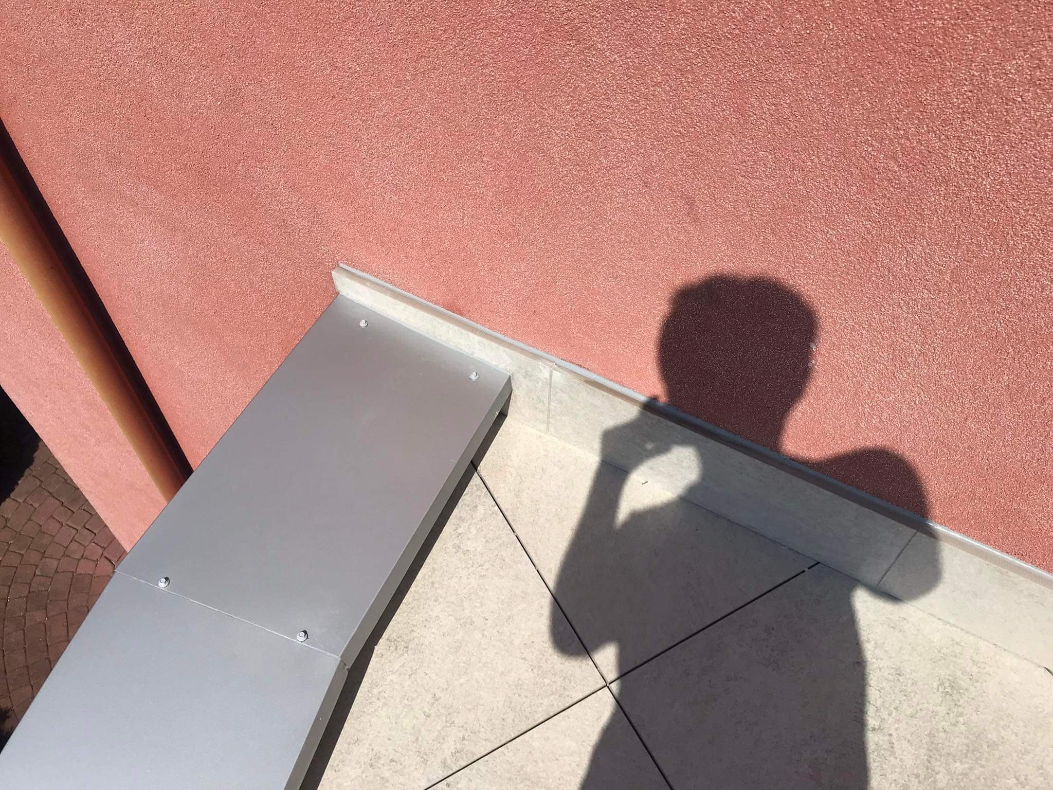 Fragment betonowego tarasu z jasnymi płytkami, widoczny cień osoby robiącej zdjęcie i fragment elewacji o różowej strukturze.