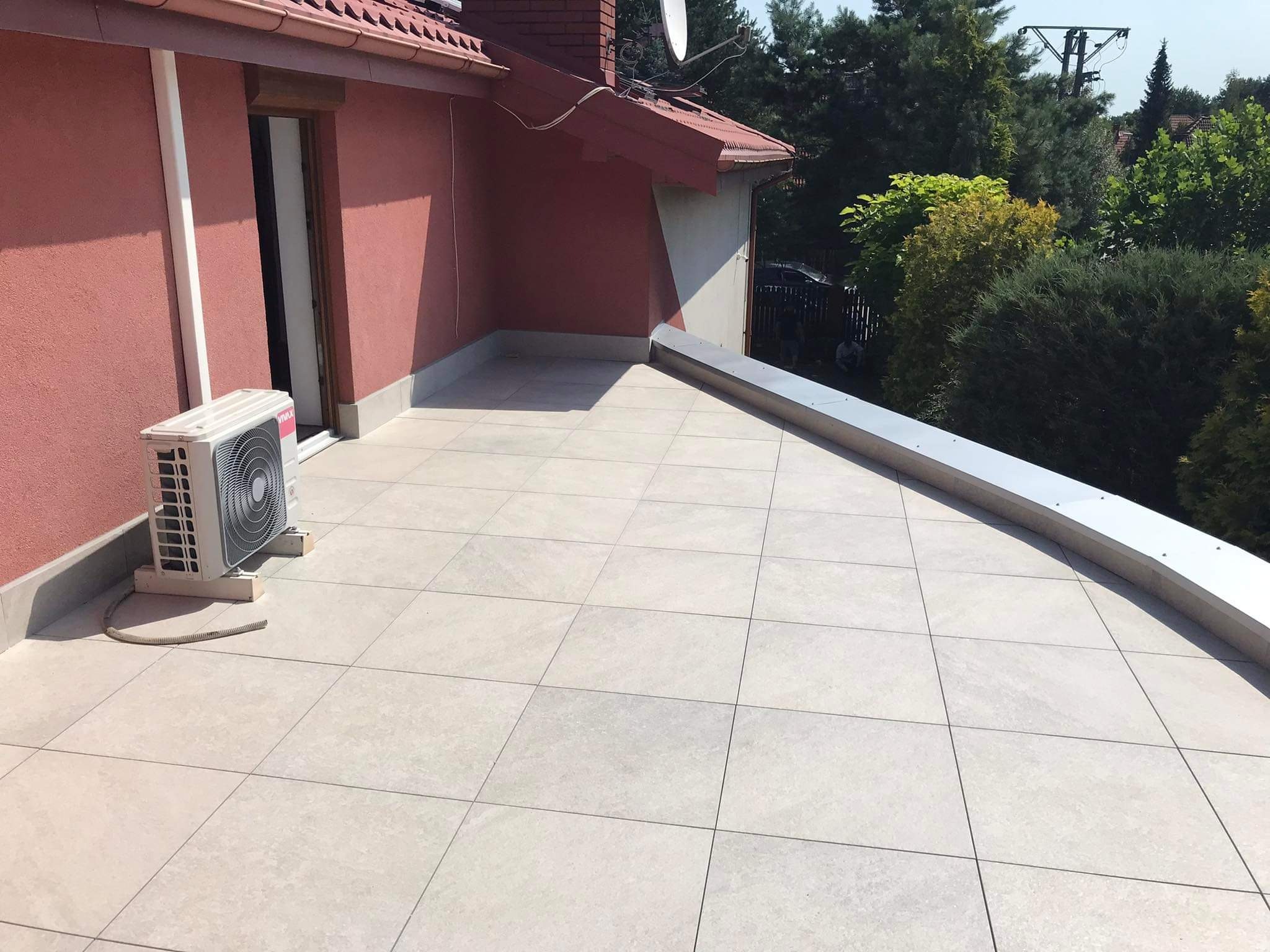 Wykończony taras z jasnych płytek, z widocznym klimatyzatorem przy ścianie budynku o różowej elewacji i metalową obróbką blacharską na krawędzi, otoczony zielenią.