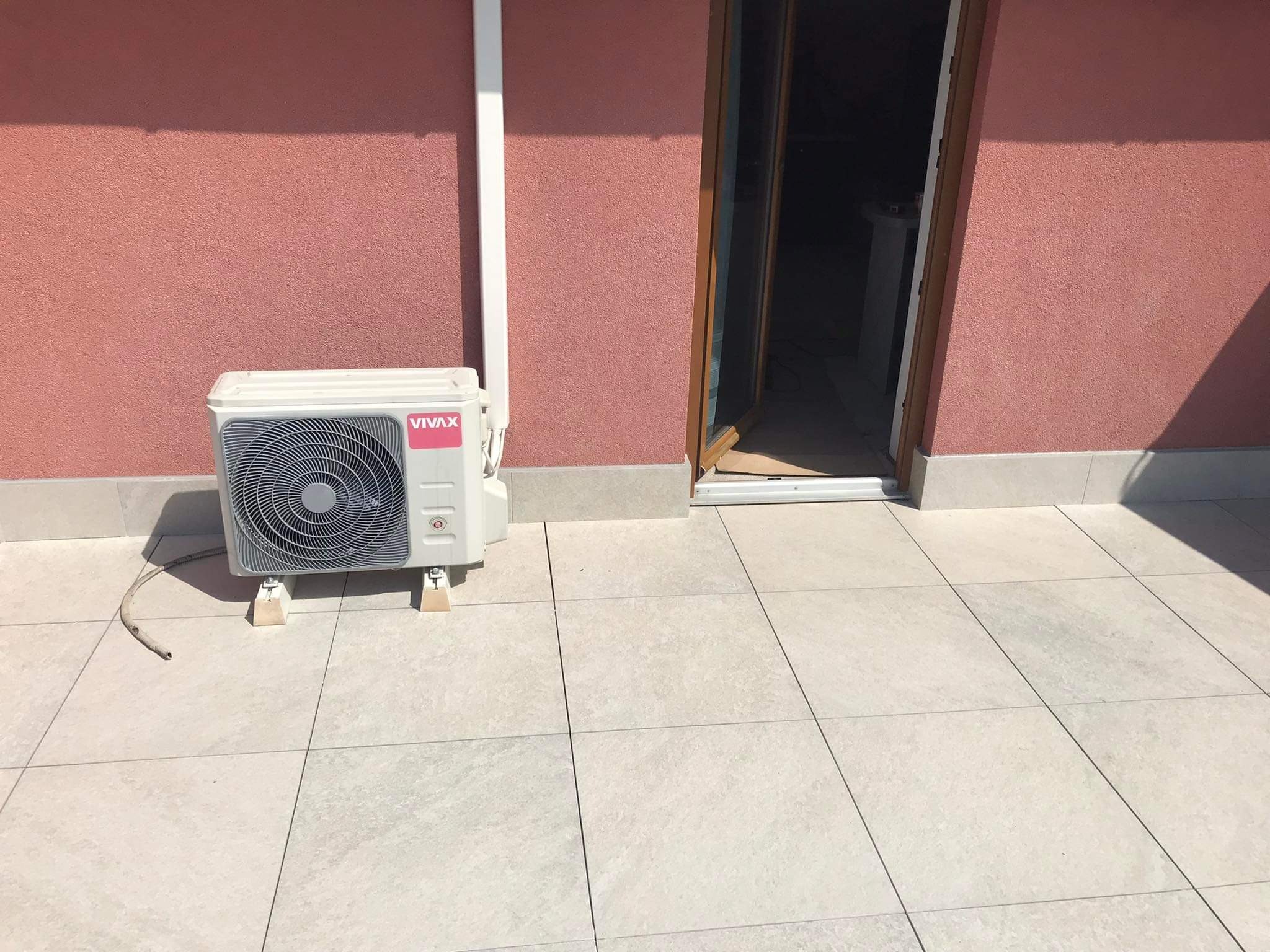 Klimatyzator marki Vivax zamontowany na podwyższeniach na wyłożonym płytkami tarasie, ściana budynku pomalowana na terakotowy kolor, otwarte drzwi balkonowe.