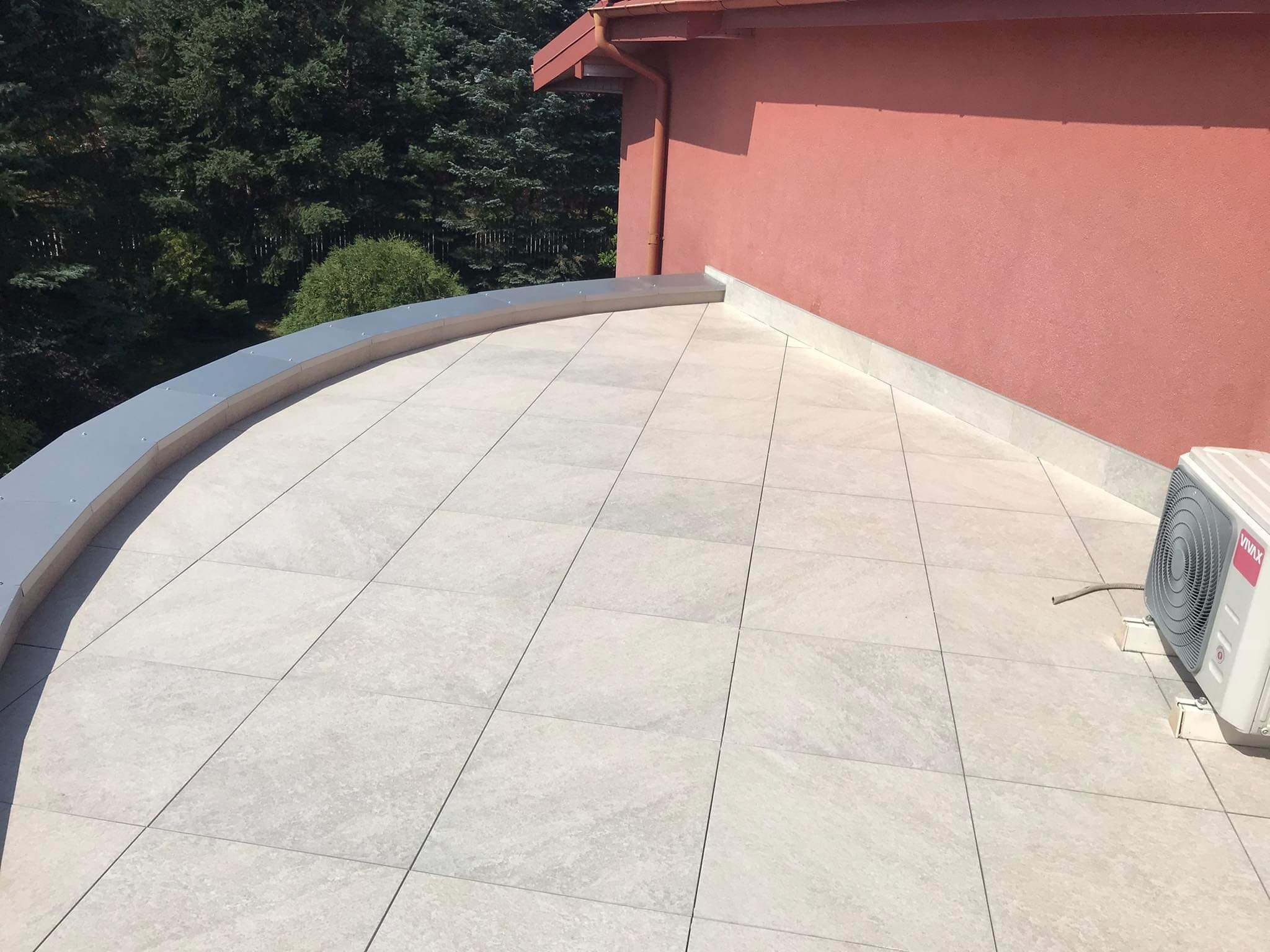 Wykończony taras na dachu budynku z jasnoszarych płyt betonowych, z widocznym obrzeżem z blachy i klimatyzatorem przy ścianie budynku.