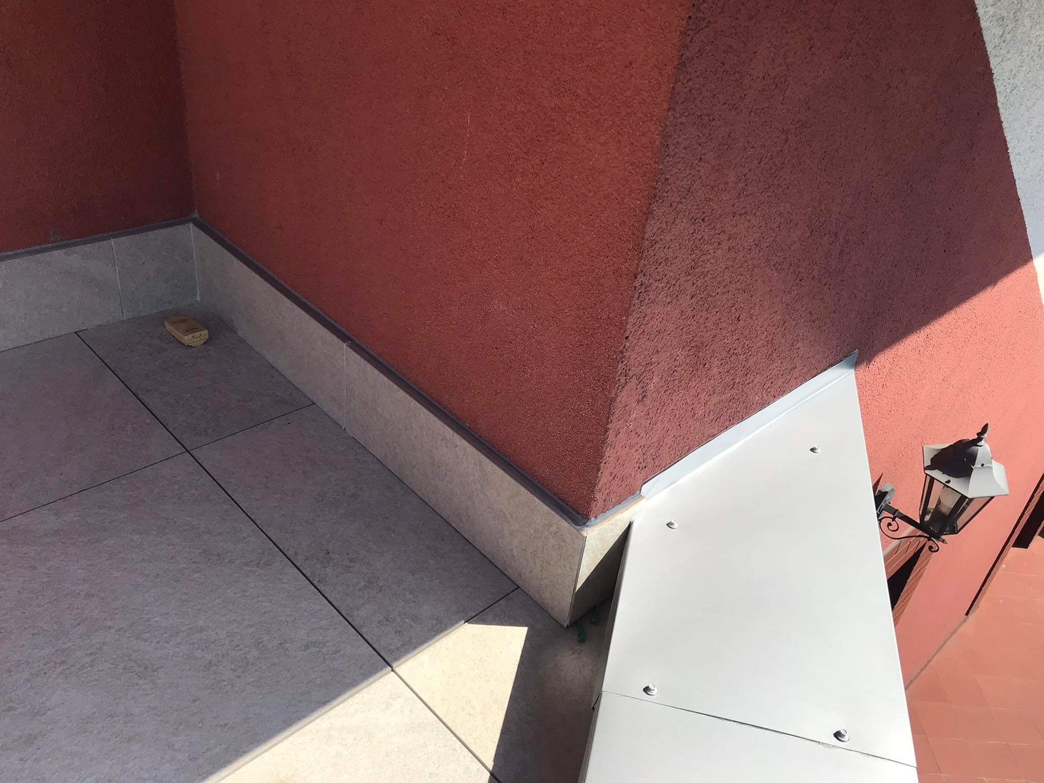 Wykończony narożnik tarasu betonowego z szarymi płytkami, widoczny fragment elewacji w kolorze ceglastym i biała balustrada z lampą.