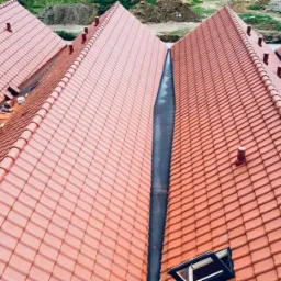 Montaż czerwonej dach&oacute;wki ceramicznej na kilku połączonych dachach dwuspadowych, widok z g&oacute;ry na dekarza pracującego na jednym z dach&oacute;w.
