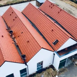 Montaż rynien na dachu z czerwonej dachówki karpiówki, widok z góry, pracownik w trakcie instalacji, budynek z białymi ścianami, otoczenie w trakcie zagospodarowywania.