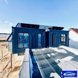 Nowoczesny dom w trakcie budowy z grafitową blachą na dachu, okna zabezpieczone folią, widok z góry na pokrycie papą i logo firmy w rogu.