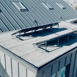 Szary dach z blachy trapezowej z oknami dachowymi i konstrukcją wsporczą, widok z góry, w tle zabudowa miejska, w prawym dolnym rogu logo firmy.