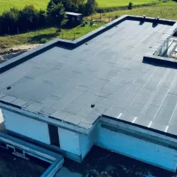 Nowoczesny dom w trakcie budowy z płaskim dachem pokrytym papą termozgrzewalną, widok z góry na tle zielonego krajobrazu.