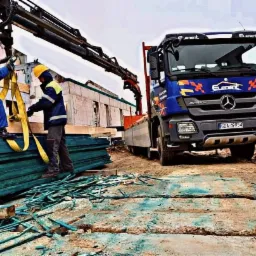 Dwóch pracowników w kaskach zabezpiecza za pomocą pasów transportowych pakiet impregnowanego drewna budowlanego na tle ciężarówki z dźwigiem i wznoszonego budynku w stanie surowym otwartym.