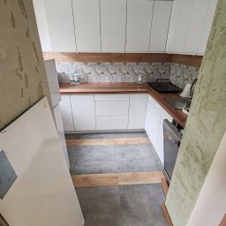 PikulBud - Nowoczesna, jasna kuchnia z białymi szafkami i drewnianym blatem. Uwagę przykuwają płytki nad blatem z motywem liści i oryginalna podłoga z szarych i drewnianych paneli.