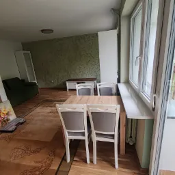 Jasne, przestronne wnętrze z drewnianym stołem i krzesłami. Dekoracyjna ściana w tle, okno z widokiem na zieleń. Na podłodze dywan w orientalne wzory i obrazy oparte o ścianę.