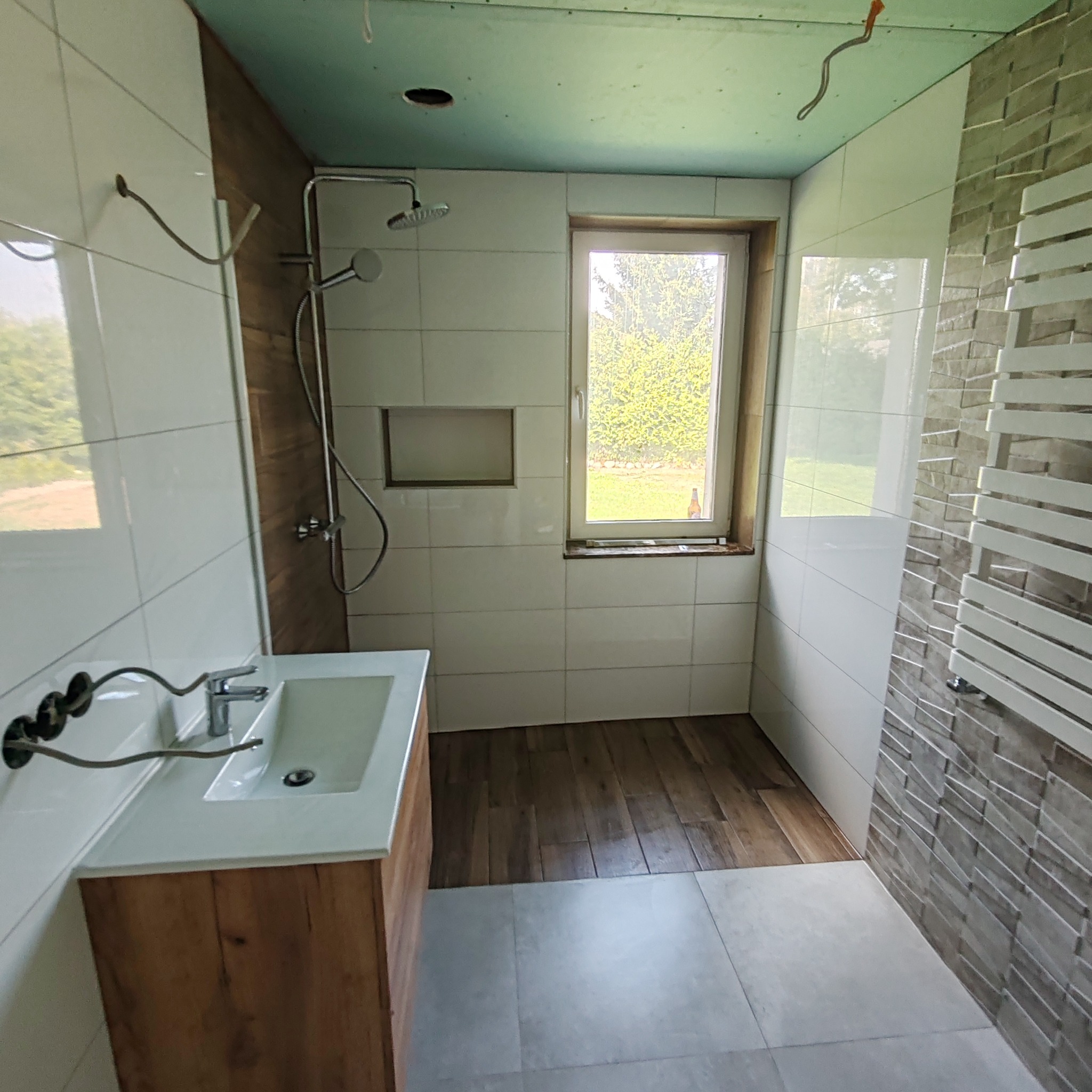 Wnętrze łazienki w trakcie prac wykończeniowych: białe płytki ścienne, częściowo ułożona podłoga z płytek imitujących drewno, zamontowana umywalka z szafką z okleiną drewnopodobną, widoczne...