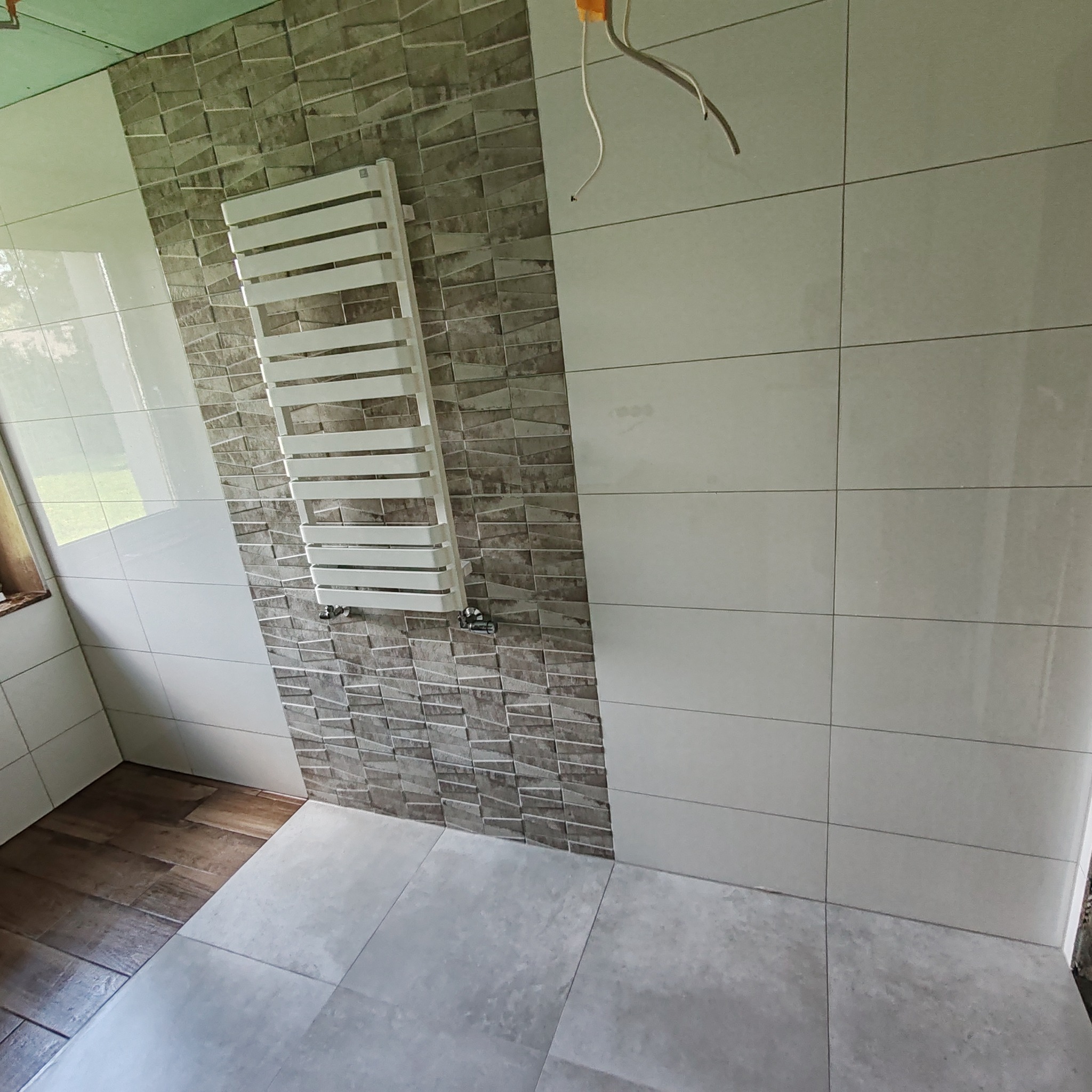 Wykończona łazienka z grzejnikiem drabinkowym na ścianie wyłożonej mozaiką w odcieniach szarości, obok ściana z jasnych, prostokątnych płytek oraz szare płytki podłogowe. Widoczne wystające kable...