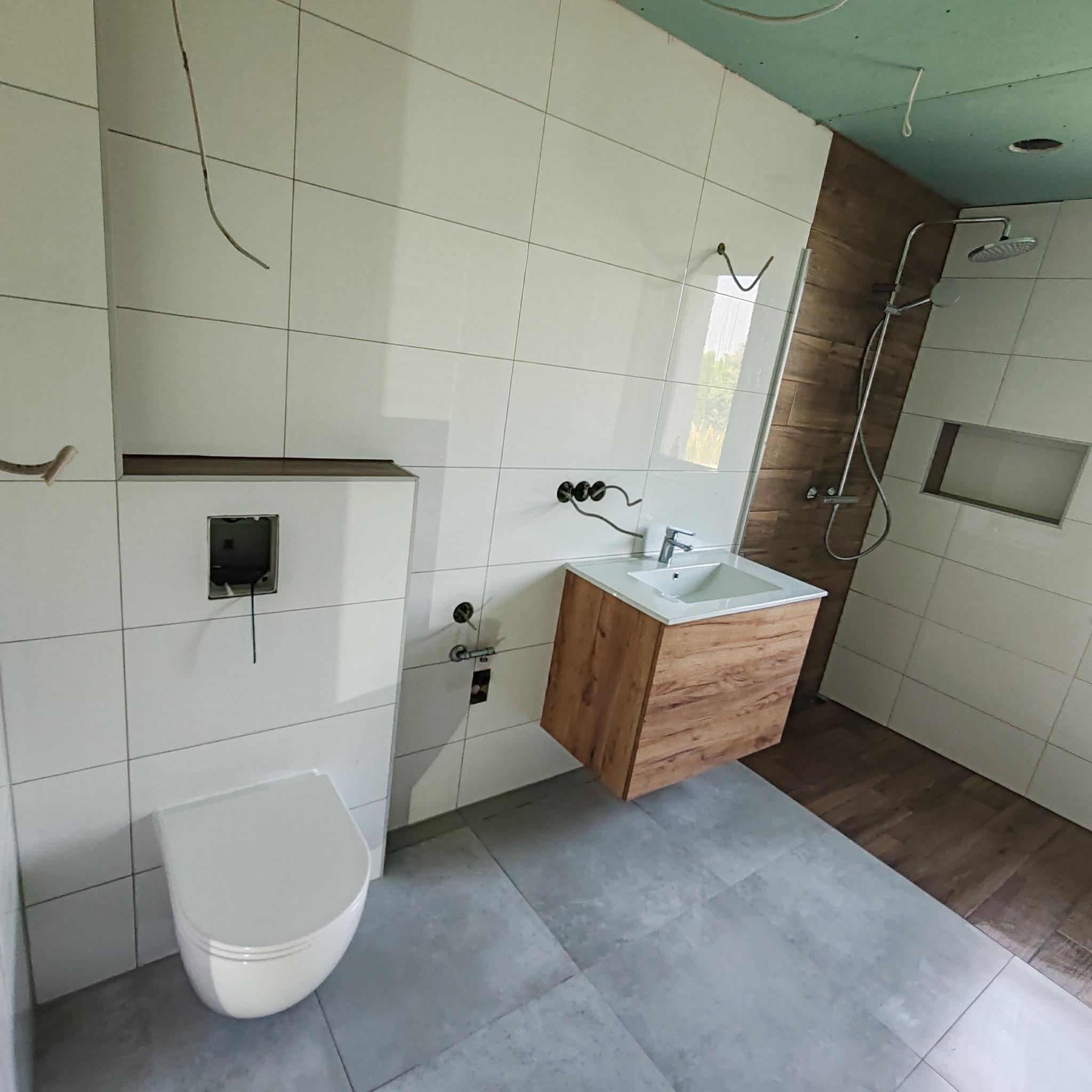 Wnętrze łazienki w trakcie prac w Lublinie: białe płytki ścienne, szare płytki podłogowe, podwieszana toaleta z instalacją w ścianie, drewniana szafka pod umywalką, widoczne przewody elektryczne.