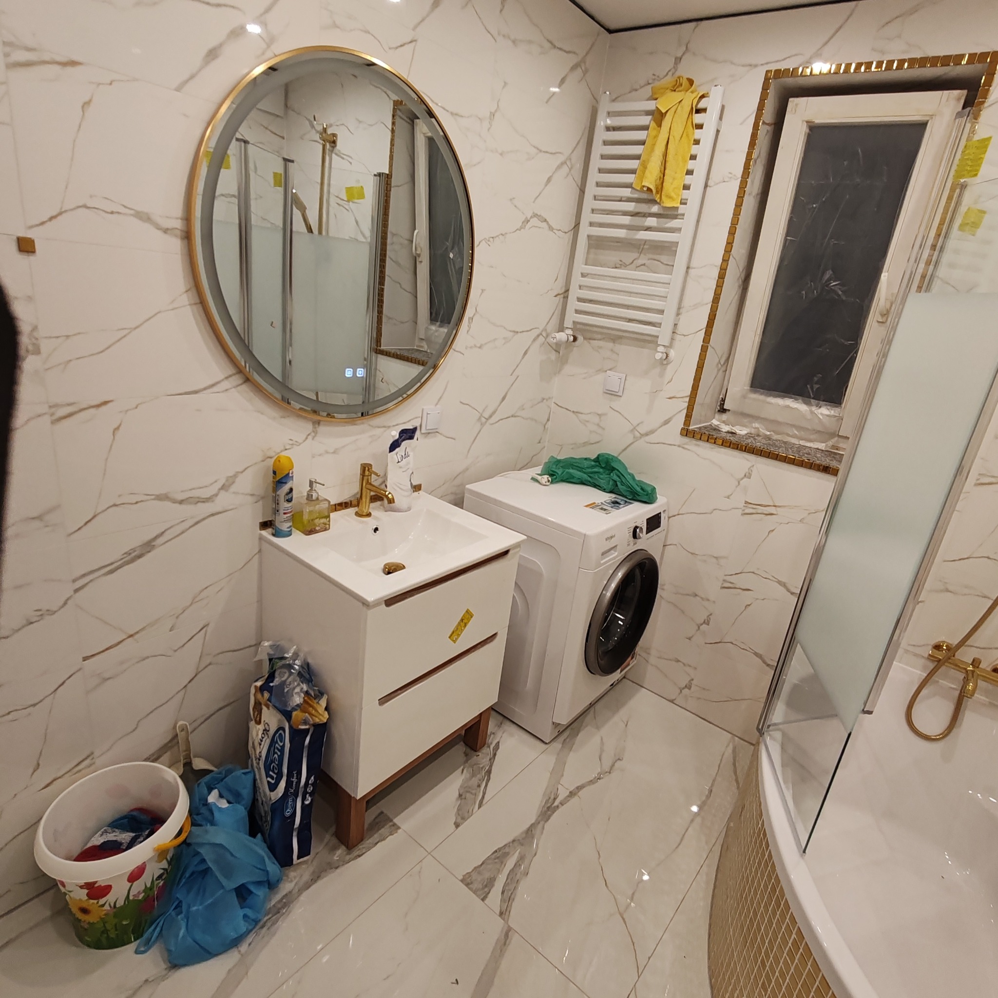 Wykończona łazienka z marmurowymi płytkami, okrągłym lustrem w złotej ramie, białą szafką umywalkową z drewnianymi nogami, pralką i kabiną prysznicową z złotą armaturą. Widoczne akcesoria...