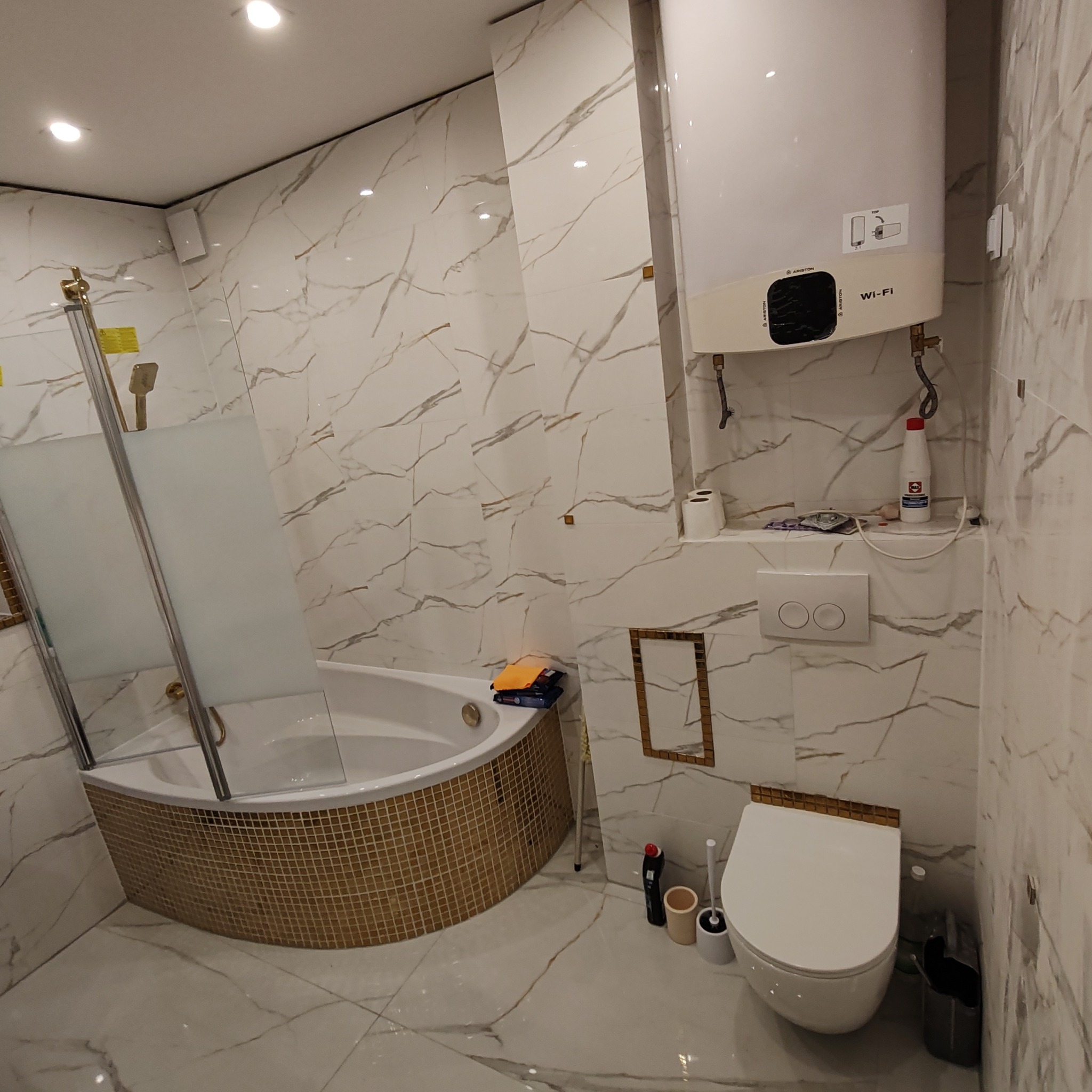 Wykończona łazienka z wanną obudowaną mozaiką, kabiną prysznicową ze złotymi elementami, wiszącą toaletą oraz białymi marmurkowymi płytkami na ścianach i podłodze, uzupełniona o bojler Ariston.