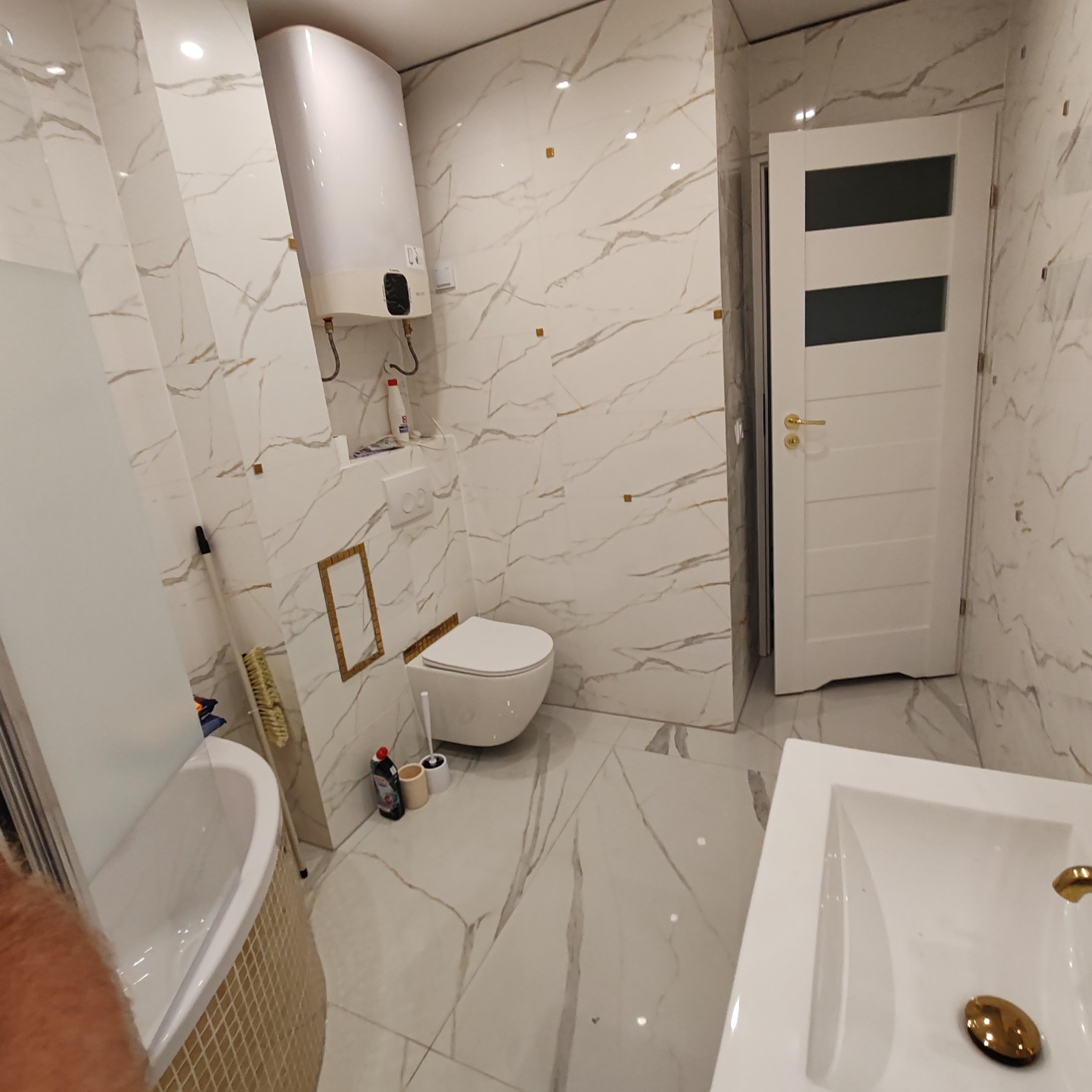 Nowoczesna łazienka z białymi płytkami imitującymi marmur, wiszącą toaletą, grzejnikiem, białą umywalką z złotą armaturą i drzwiami z przeszkleniami.