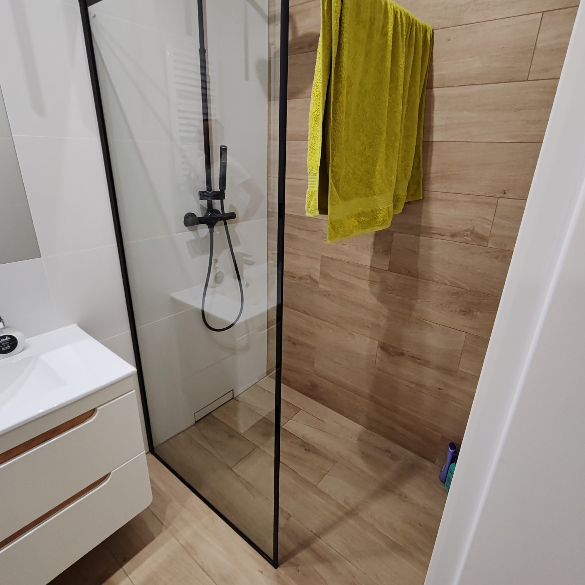 Nowoczesna łazienka z kabiną prysznicową typu walk-in z czarnymi profilami, jasnymi płytkami imitującymi drewno na ścianie i podłodze, oraz białymi płytkami na ścianie, z żółtym ręcznikiem wiszącym...