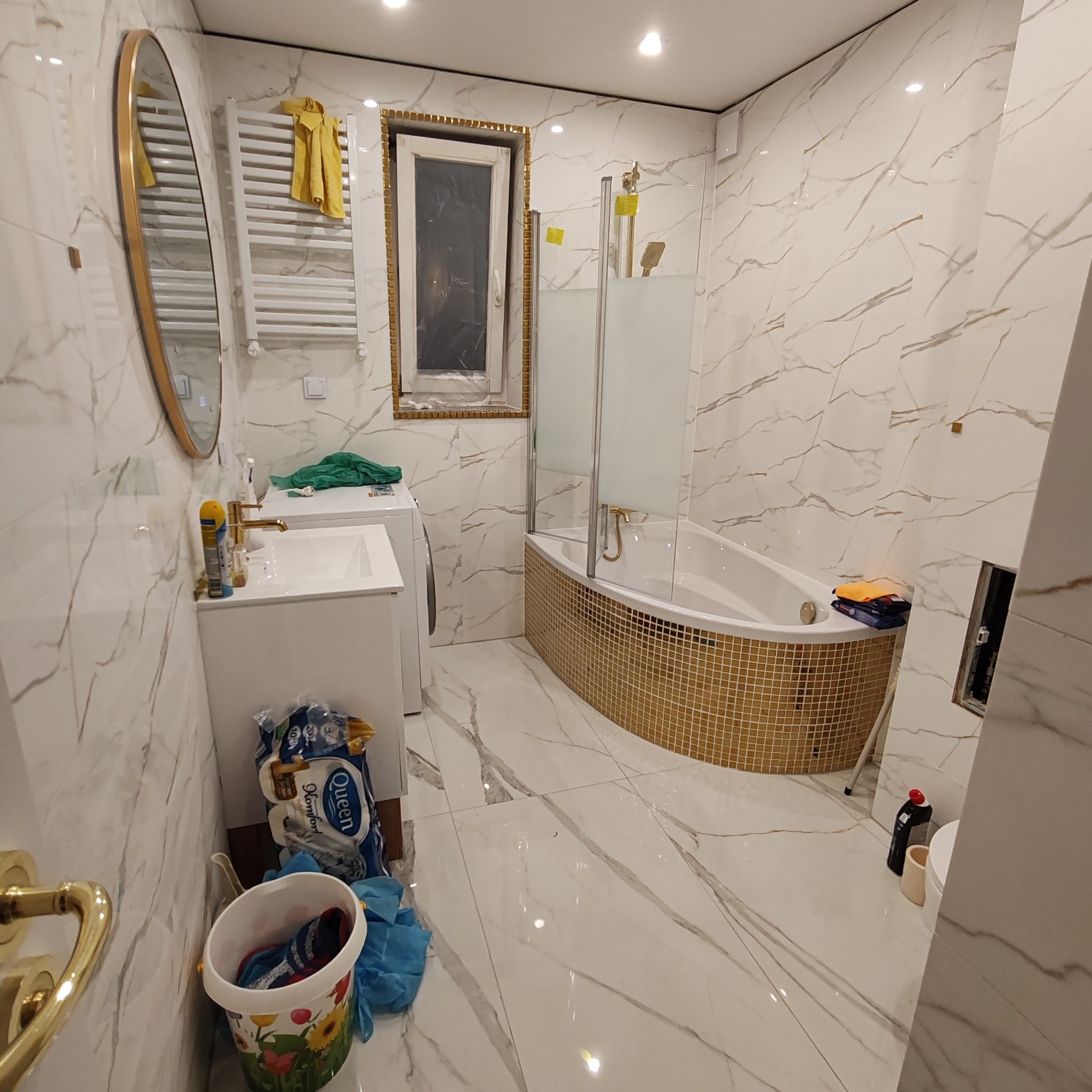 Wykończona łazienka z wanną obudowaną złotą mozaiką, białe płytki imitujące marmur na ścianach i podłodze, okrągłe lustro w złotej ramie i grzejnik drabinkowy. Umywalka z szafką i pralka w rogu...