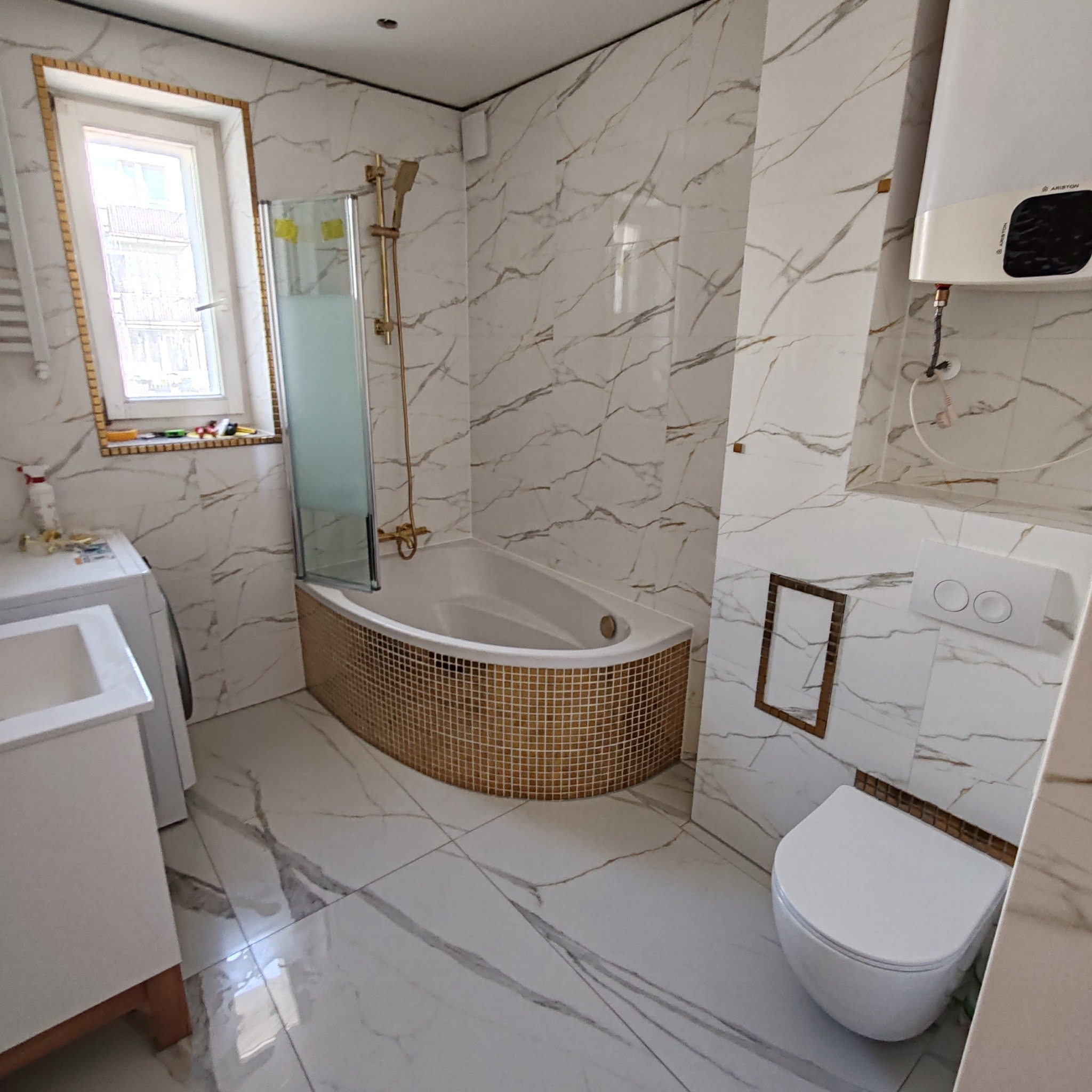 Wykończona łazienka z wanną obudowaną mozaiką, białe marmurowe płytki ścienne i podłogowe, złota armatura, okno z mozaikową ramą, zabudowana toaleta.