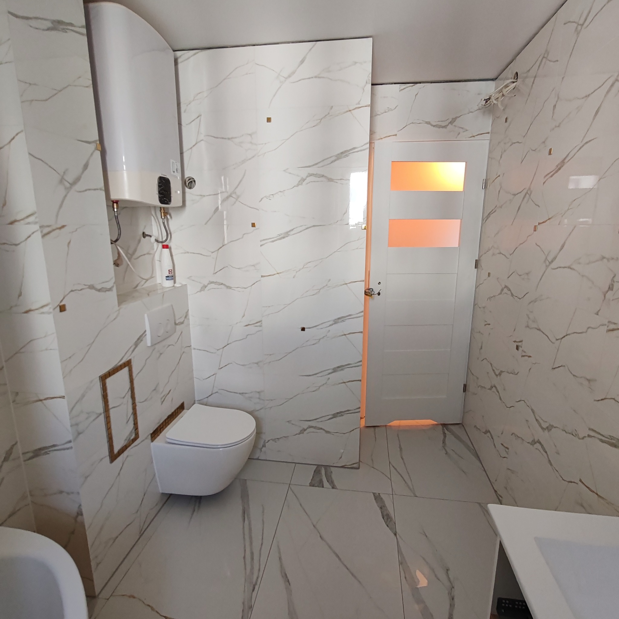 Nowoczesna łazienka z białymi płytkami imitującymi marmur, podwieszana toaleta i bojler elektryczny, złote akcenty dekoracyjne na ścianach i drzwi z pomarańczowym przeszkleniem.