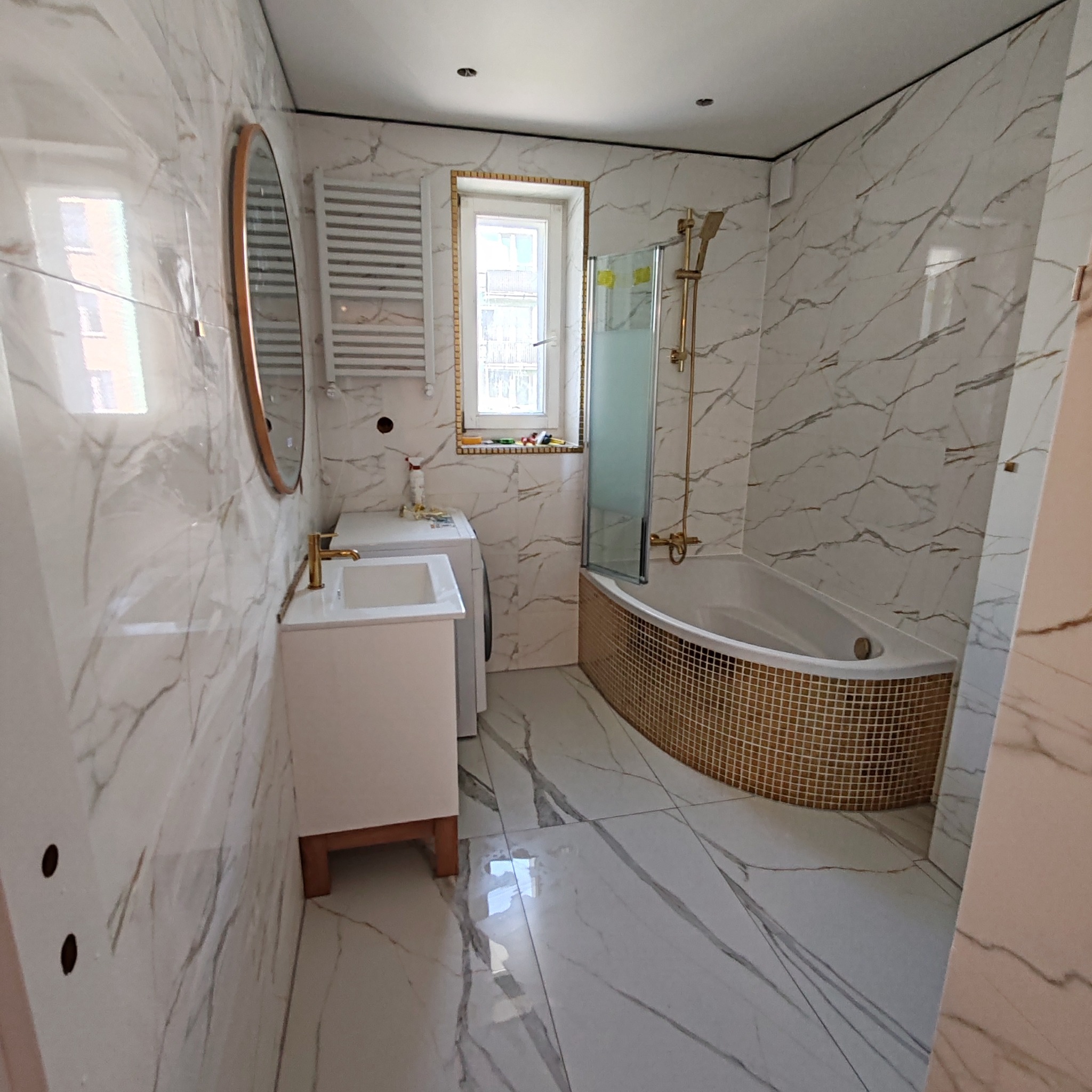 Nowoczesna łazienka z białymi marmurkowymi płytkami, mozaiką w kolorze złota na obudowie wanny, złotą armaturą, okrągłym lustrem i grzejnikiem drabinkowym, z pralką widoczną w tle.