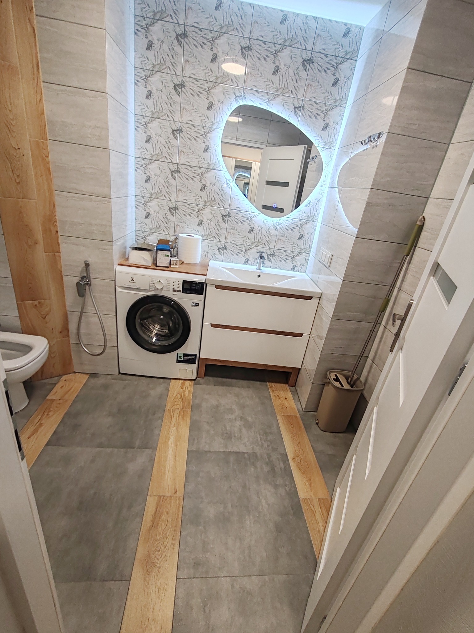 Ukończona łazienka z pralką obok umywalki i podświetlanym lustrem o nieregularnym kształcie. Na podłodze płytki imitujące drewno.