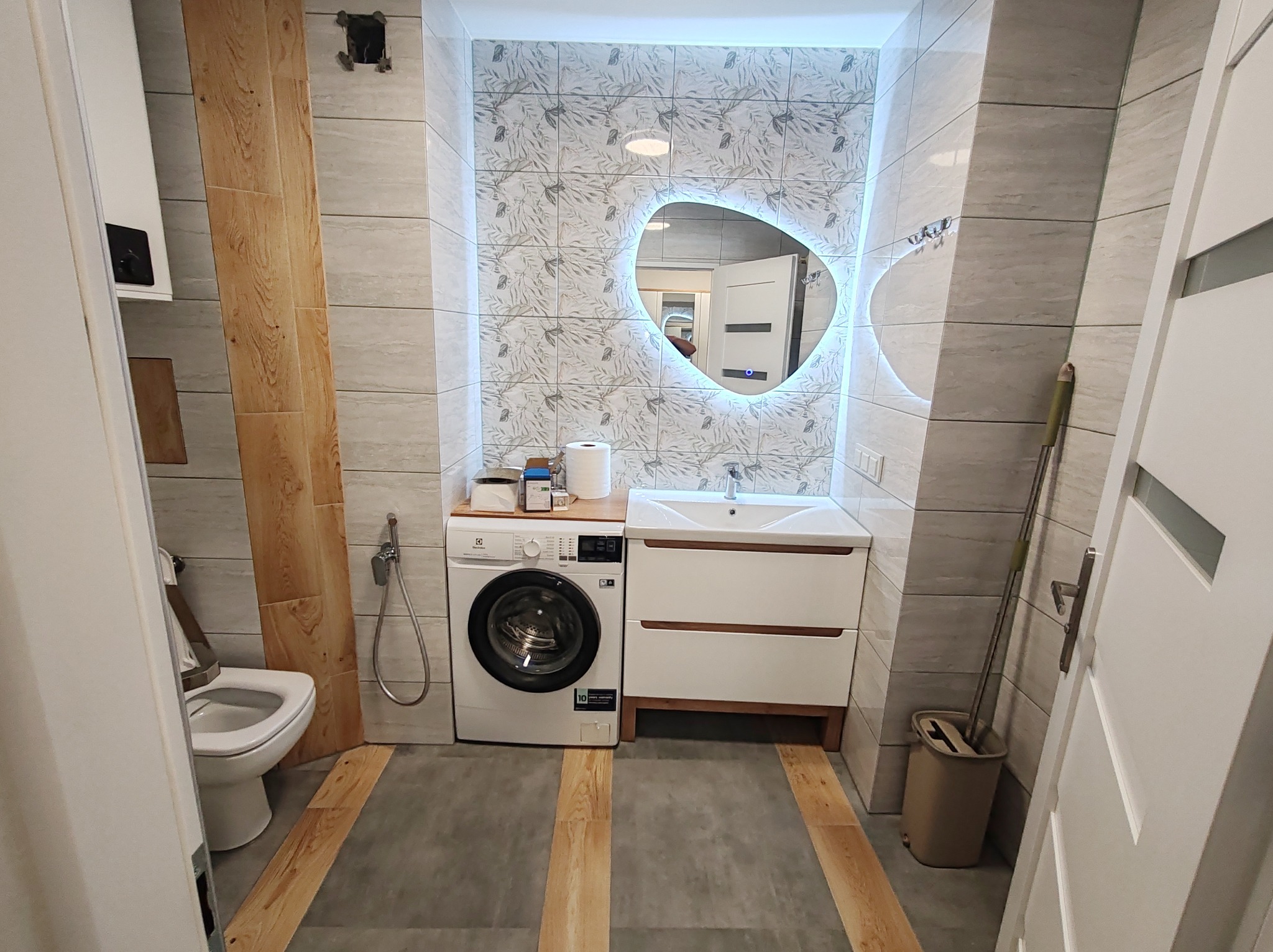 Jasna łazienka z umywalką, pralką i toaletą. Uwagę przyciąga lustro z podświetleniem LED w kształcie nieregularnego okręgu i wzorzyste płytki ścienne.