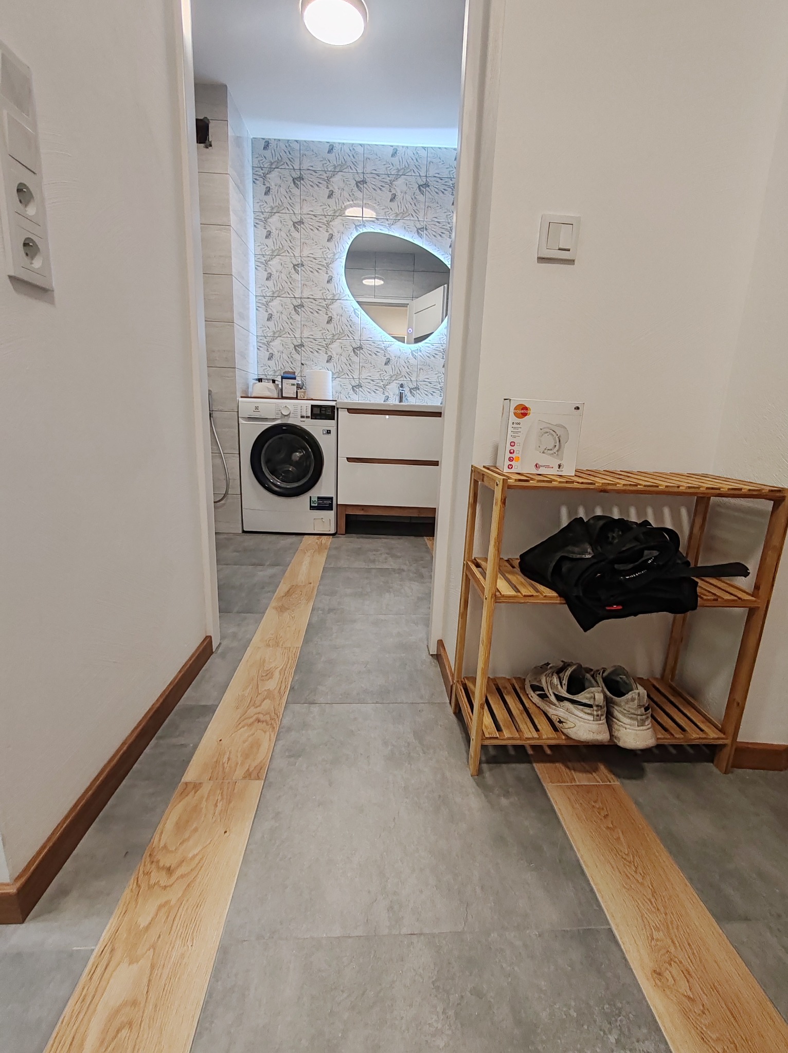 Wnętrze: szare płytki podłogowe z drewnianymi wstawkami prowadzą do łazienki z pralką i lustrem w kształcie serca. Obok drewniany regał na buty z pudełkiem grzejnika.