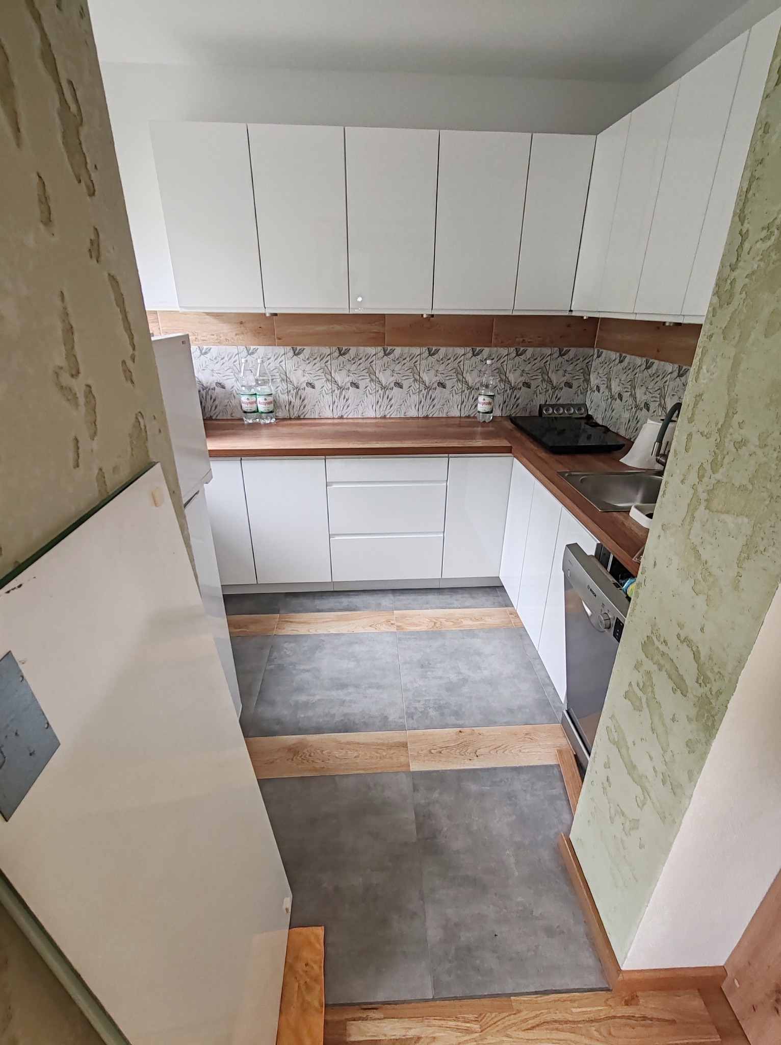 Nowoczesna, jasna kuchnia z białymi szafkami i drewnianym blatem. Uwagę przykuwają płytki nad blatem z motywem liści i oryginalna podłoga z szarych i drewnianych paneli.