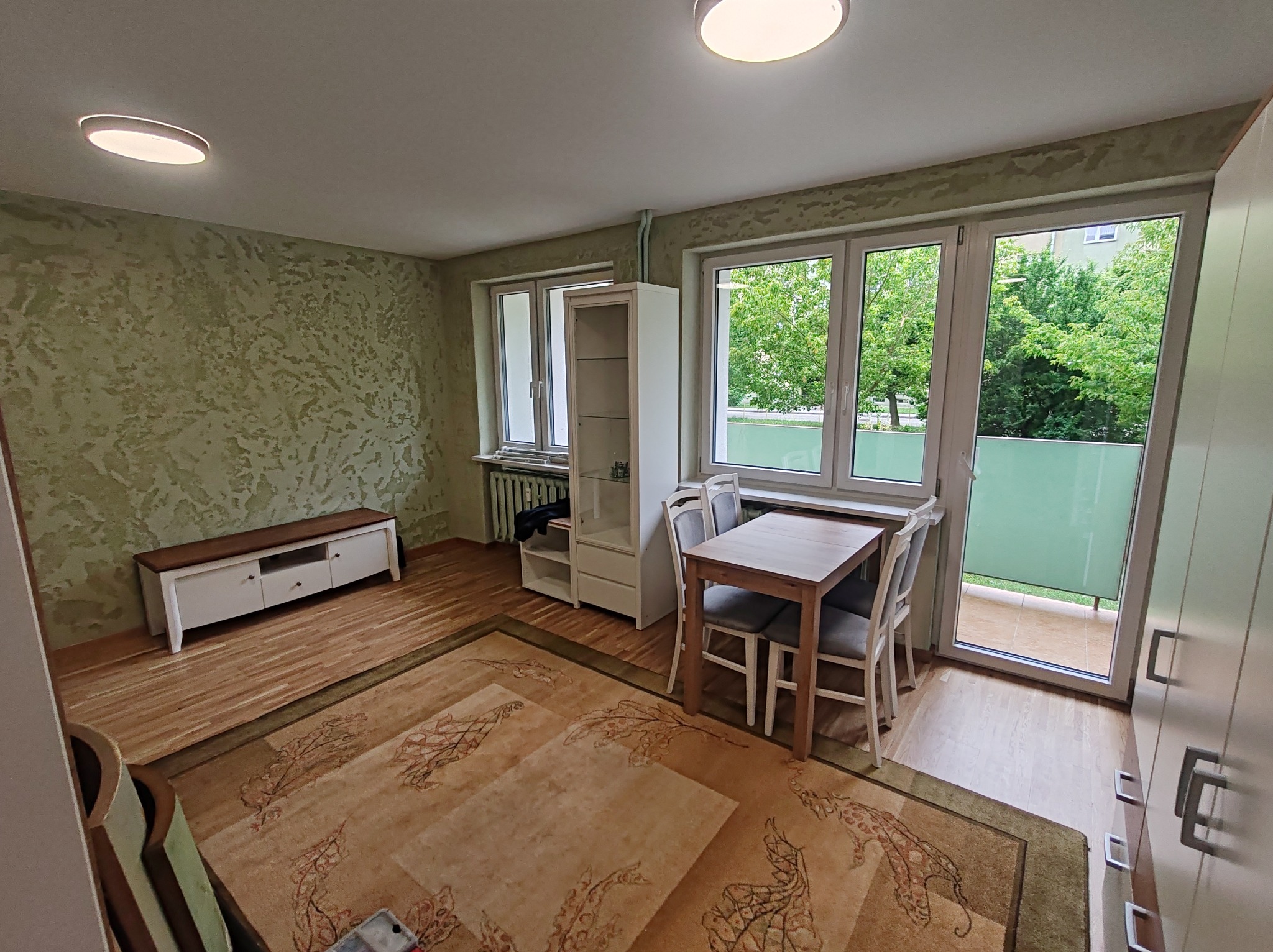 Jasne wnętrze pokoju z teksturowaną ścianą, drewnianą podłogą i dywanem w orientalne wzory. Widoczne meble: stół, krzesła, szafka RTV i regał. Za oknem zieleń.