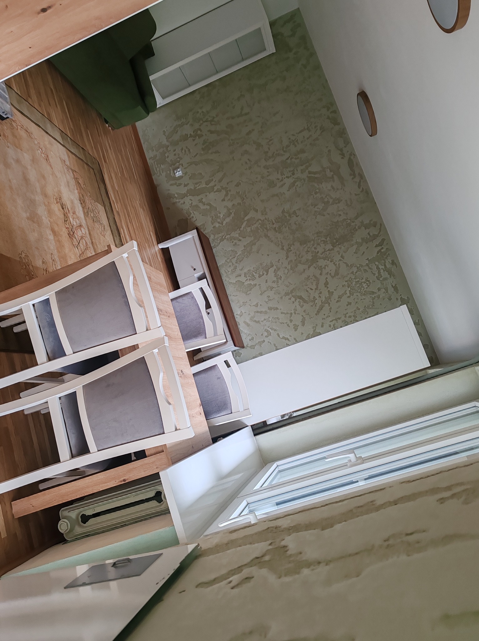 Ustawiony stół z krzesłami w jasnym wnętrzu z dekoracyjną ścianą. Widok na okno i grzejnik. Perspektywa z dołu, obrót o 90 stopni.