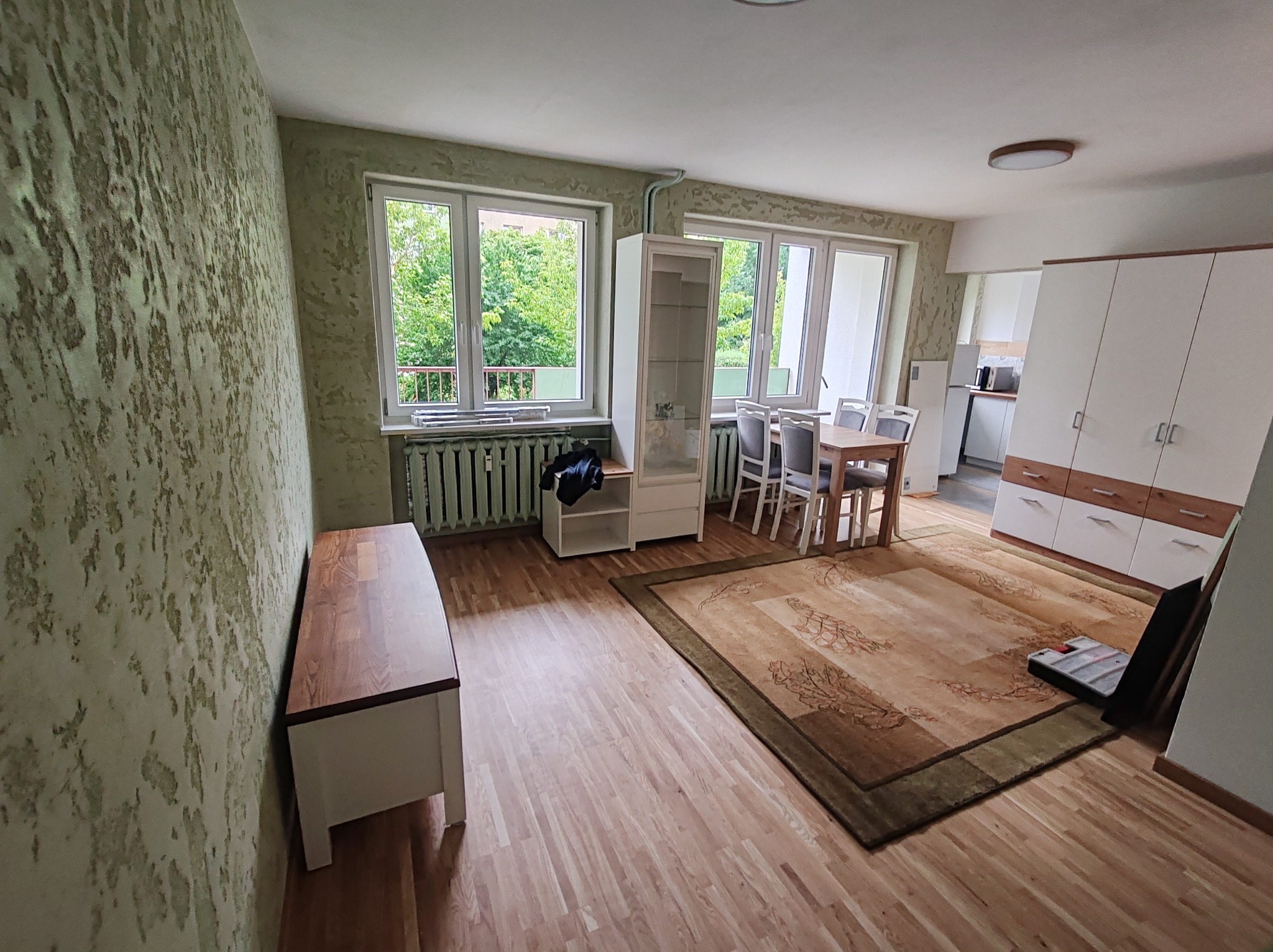 Jasne wnętrze pokoju z drewnianą podłogą, dywanem i ścianą o nieregularnej fakturze. Widoczne okna z widokiem na zieleń, stół z krzesłami i białe meble.