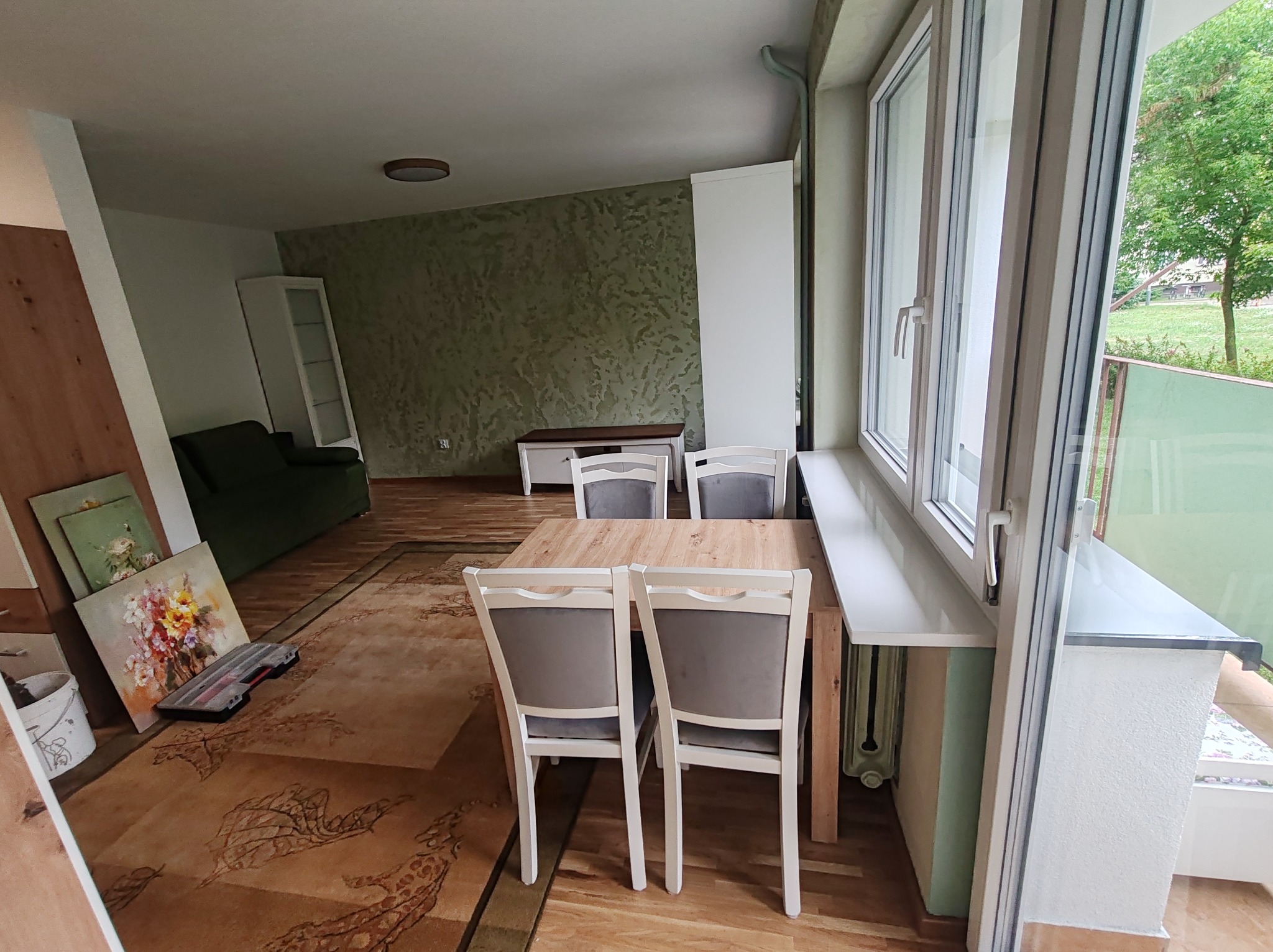 Jasne, przestronne wnętrze z drewnianym stołem i krzesłami. Dekoracyjna ściana w tle, okno z widokiem na zieleń. Na podłodze dywan w orientalne wzory i obrazy oparte o ścianę.