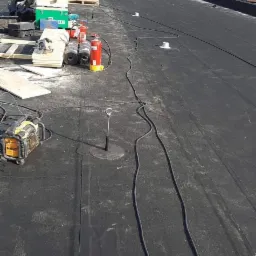 Czarny, płaski dach pokryty papą z widocznymi narzędziami, rolkami papy, butlami z gazem, oraz przewodami elektrycznymi podczas prac dekarskich w słoneczny dzień.