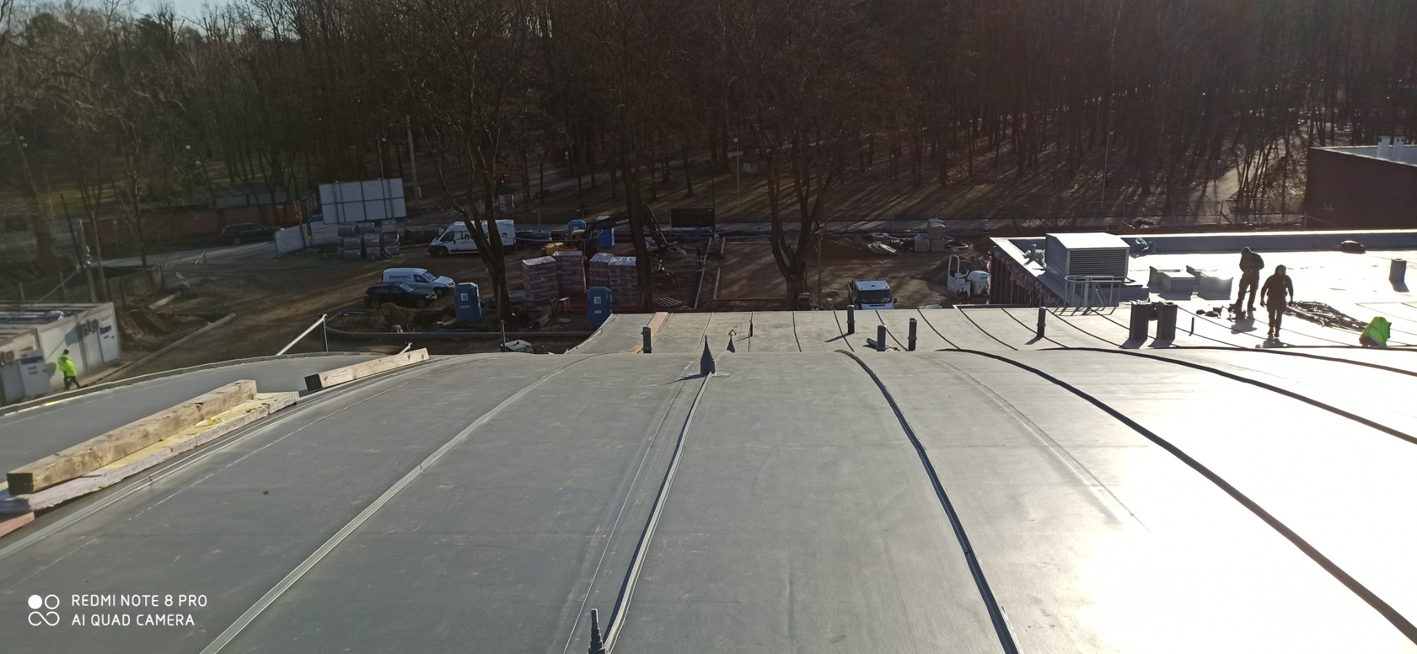 Widok z góry na duży, płaski dach pokryty szarą membraną, z widocznymi pracownikami w oddali i elementami konstrukcyjnymi na krawędzi, w tle drzewa i plac budowy.