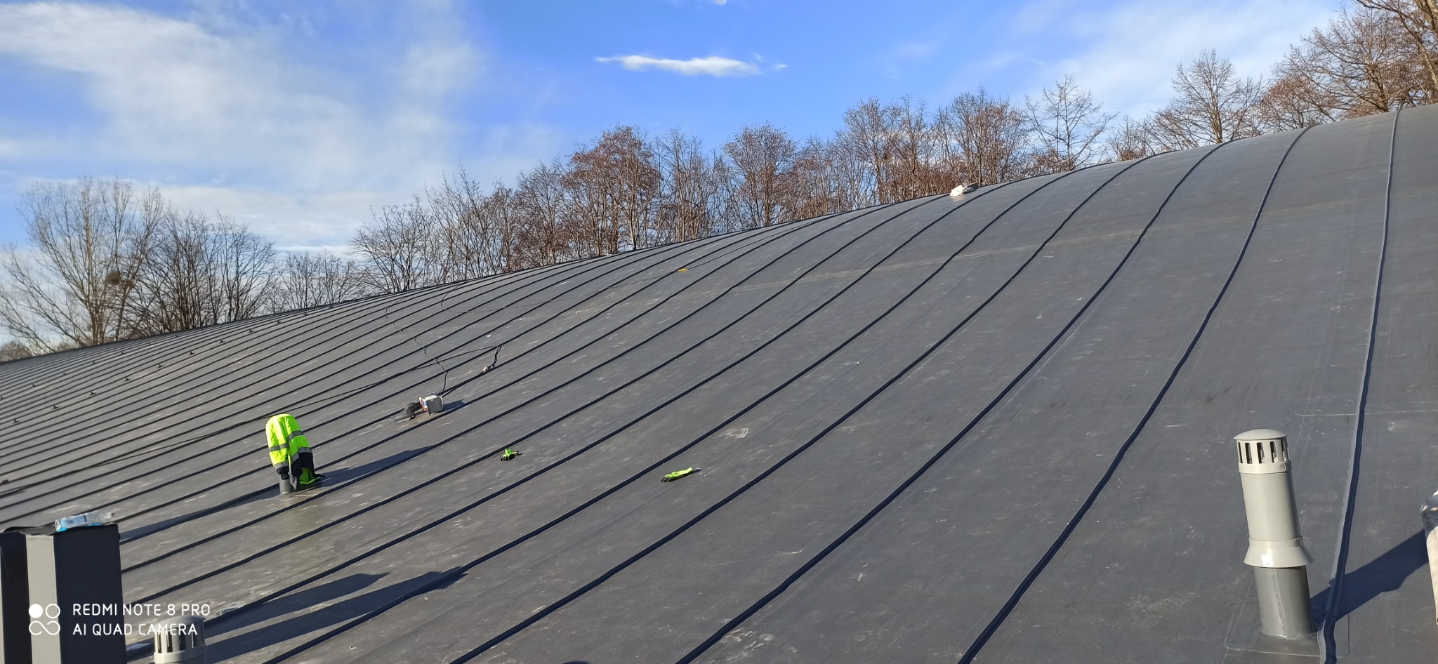 Szary, metalowy dach z widocznymi łączeniami blach, na którym stoi pracownik w odblaskowej kamizelce, w tle drzewa bez liści i błękitne niebo z chmurami.