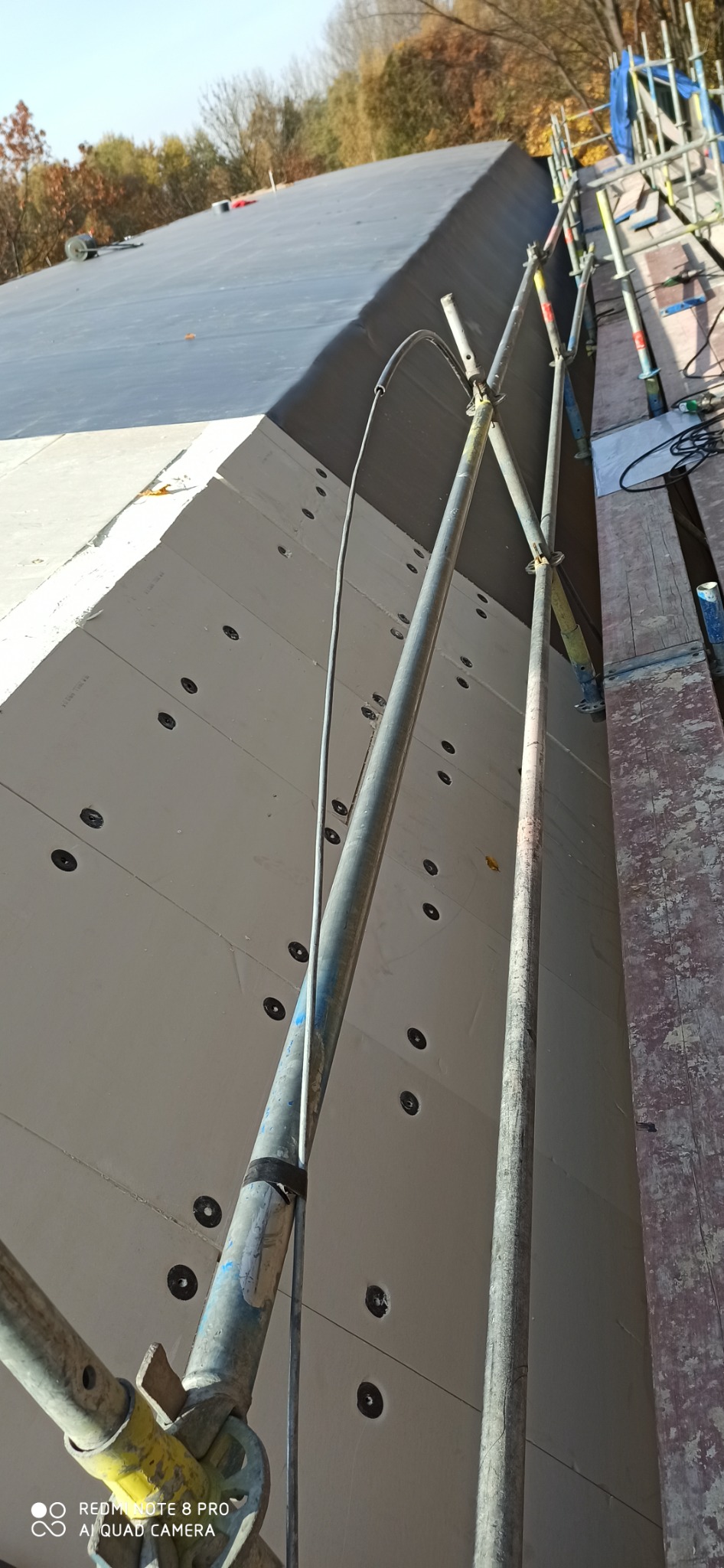 Ocieplony dach z widocznymi mocowaniami, częściowo pokryty czarną membraną, rusztowanie w trakcie prac dekarskich.