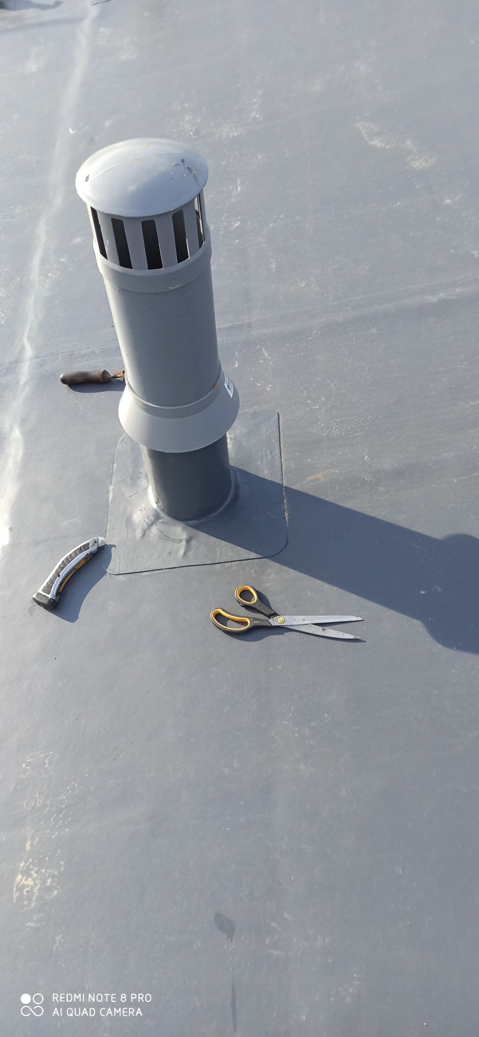 Szary, płaski dach z wentylacją kominową, obok leżą nożyczki z żółtymi uchwytami i nóż z drewnianą rączką.