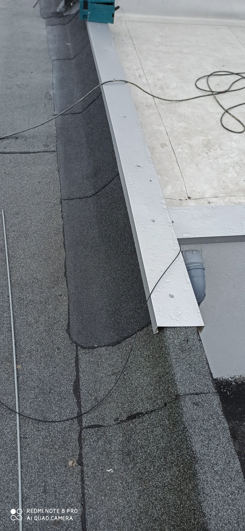 Fragment płaskiego dachu pokrytego papą z obróbką blacharską w kolorze szarym, widoczne łączenia papy i elementy odprowadzające wodę.