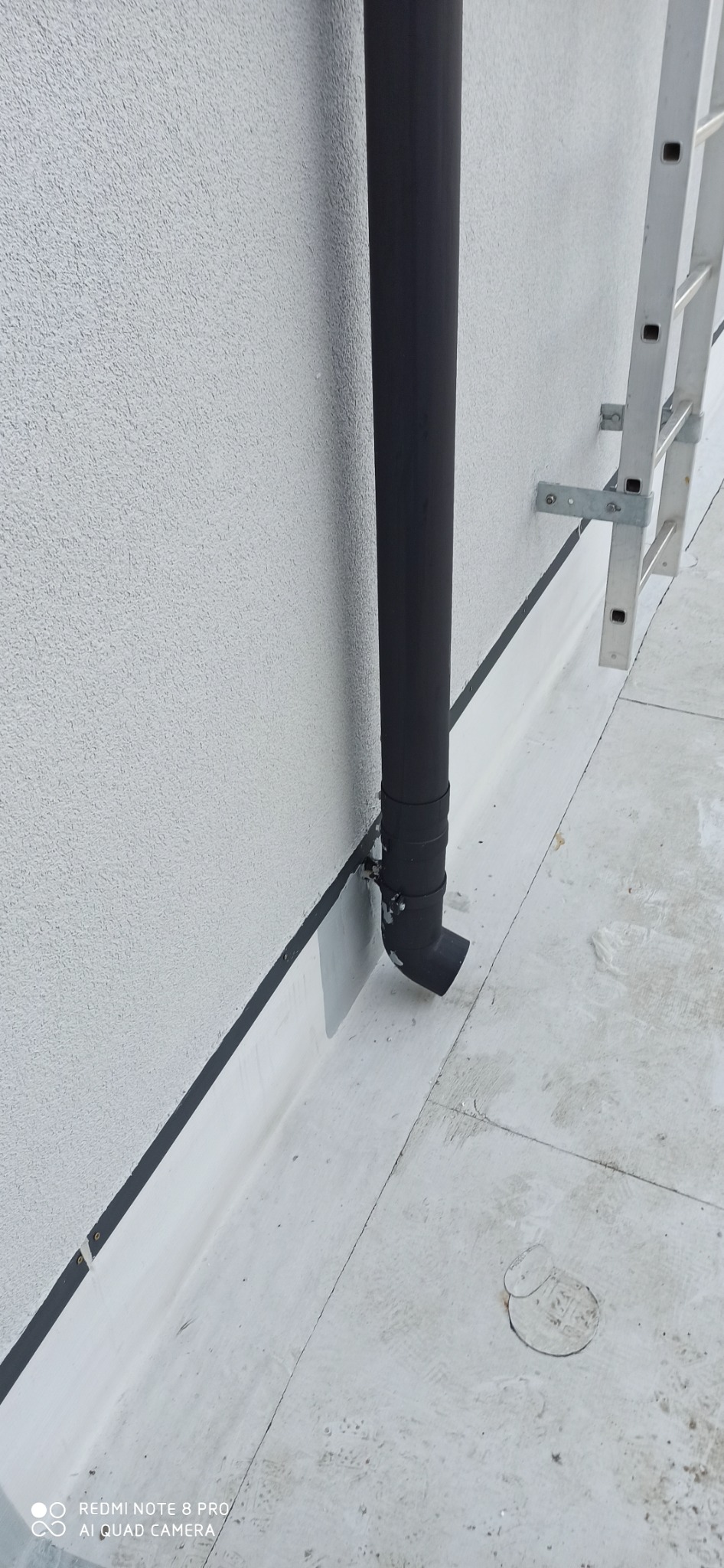 Czarne rury spustowe przymocowane do jasnej ściany budynku, z widoczną drabinką aluminiową i fragmentem dachu pokrytego papą.