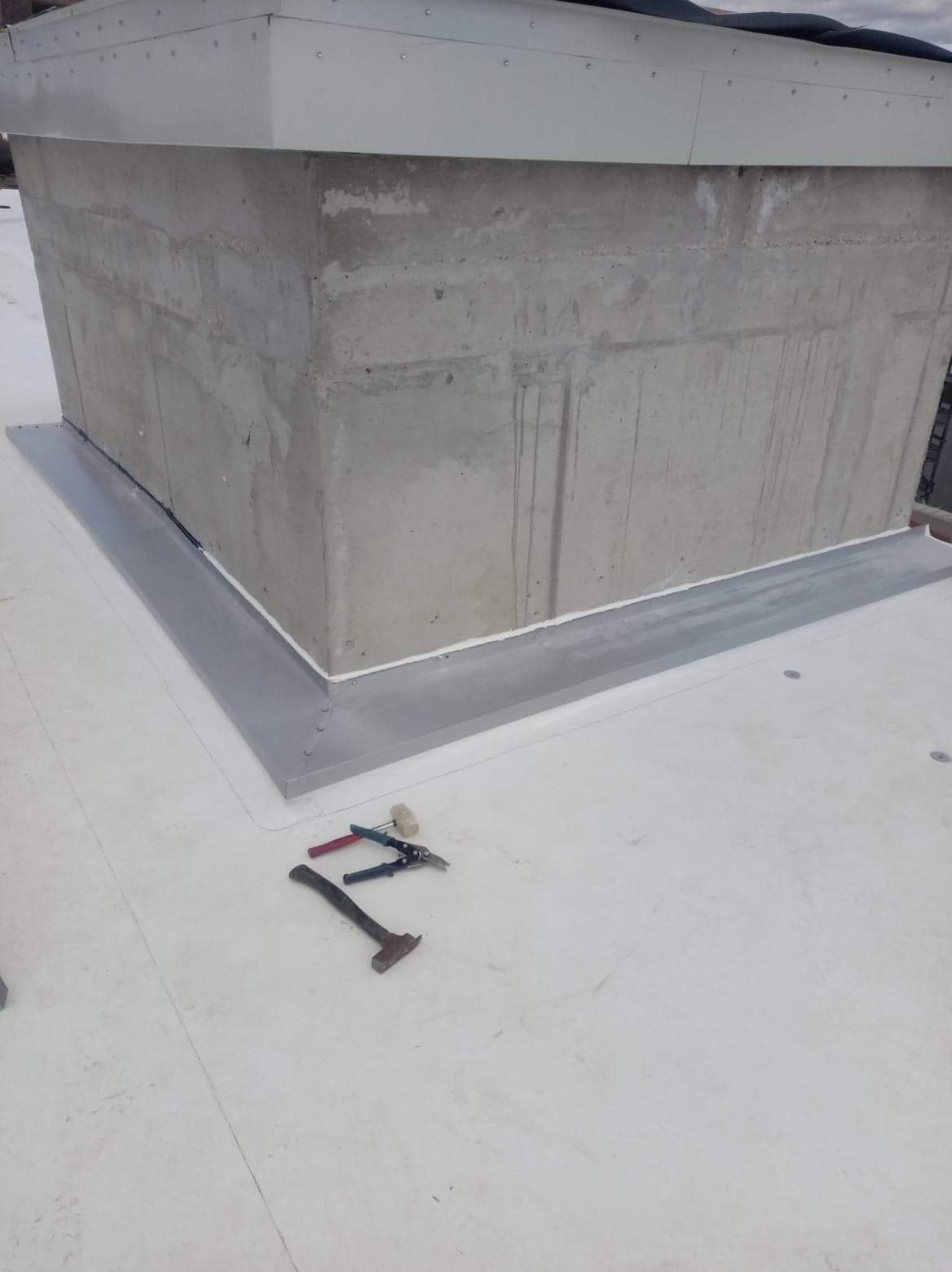 Betonowa podstawa komina na płaskim dachu z nową obróbką blacharską i narzędziami dekarskimi obok.