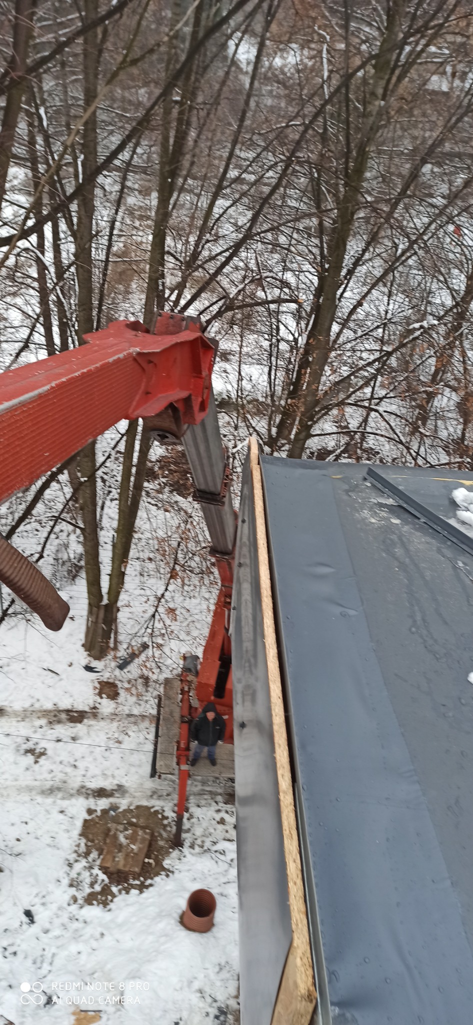 Widok z góry na fragment dachu z nową membraną, drewnianą deską i dźwigiem z operatorem na tle zimowego krajobrazu z drzewami i śniegiem.