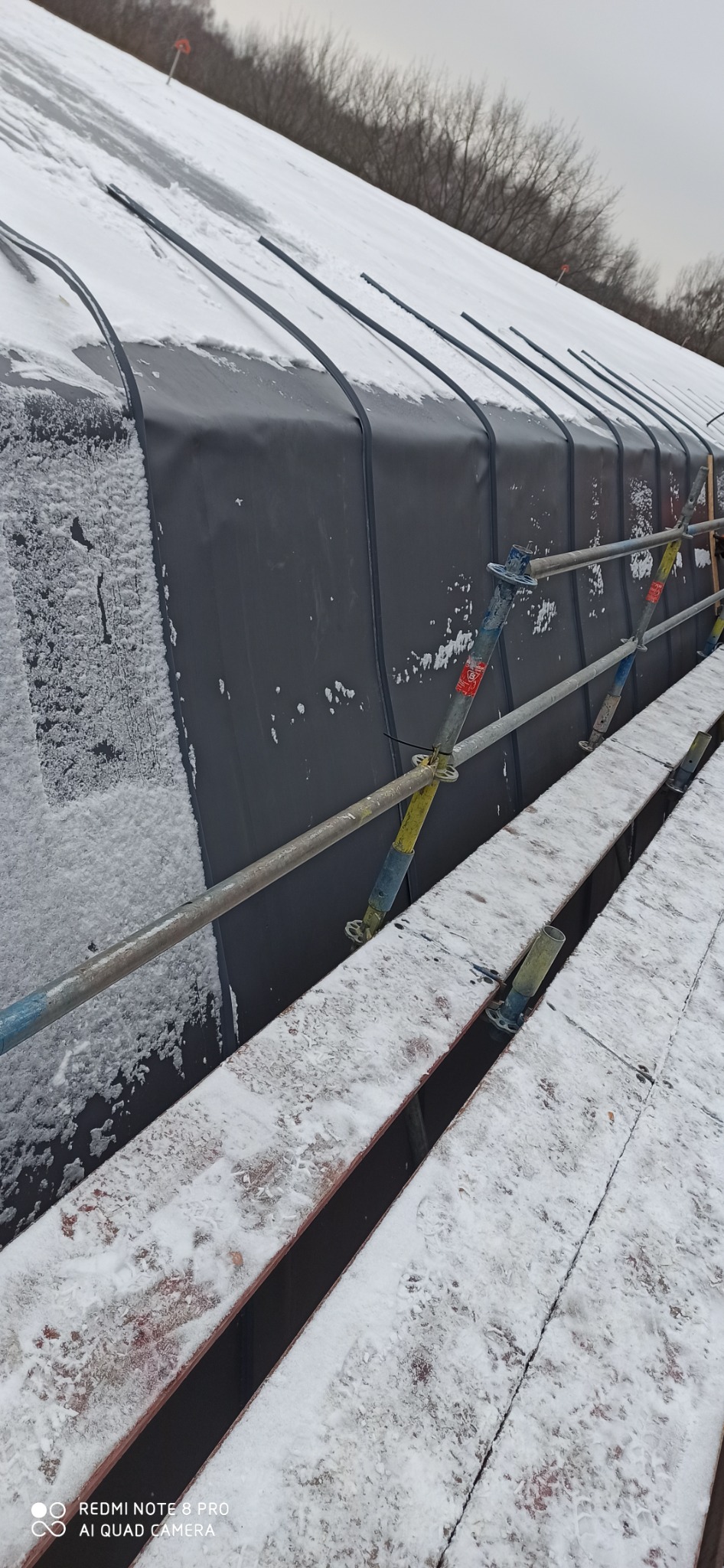 Ośnieżony dach pokryty czarną membraną dachową, widoczne elementy rusztowania i drzewa w tle.