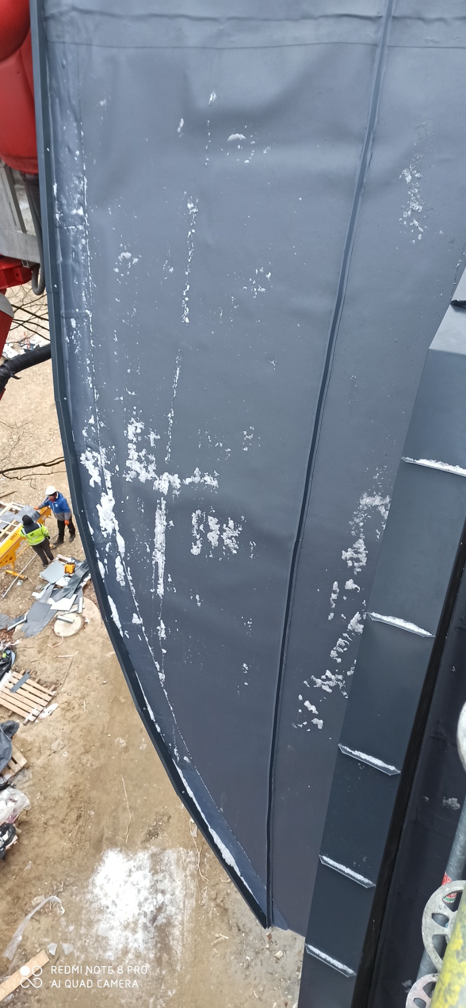 Szary, metalowy dach z widocznymi śladami białego pyłu, prawdopodobnie po cięciu, z dwoma pracownikami w oddali i elementami rusztowania w kadrze.